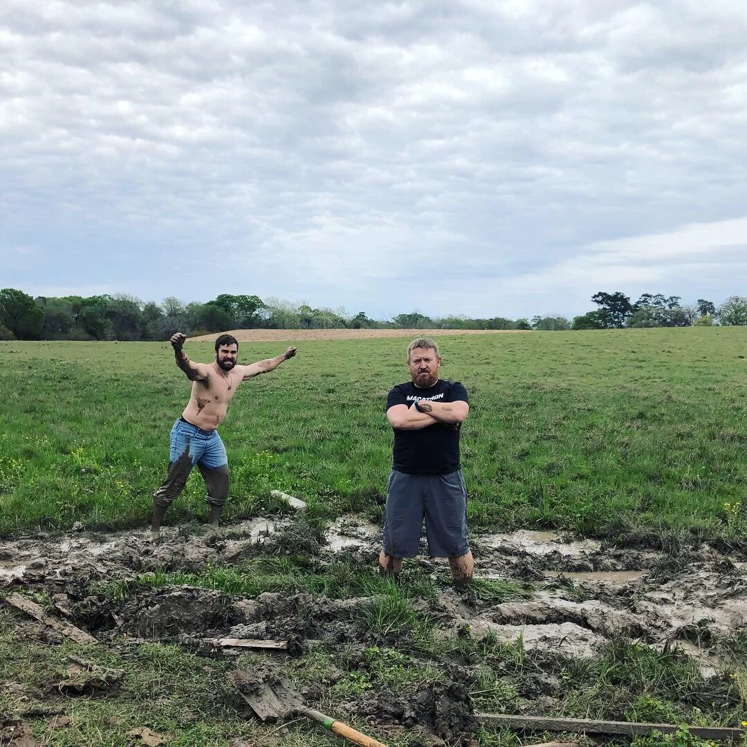 Two men standing in a muddy field, one shirtless with mud on his pants and posing with fists raised, the other with arms crossed, wearing a black t-shirt and shorts, with a green field and cloudy sky in the background.