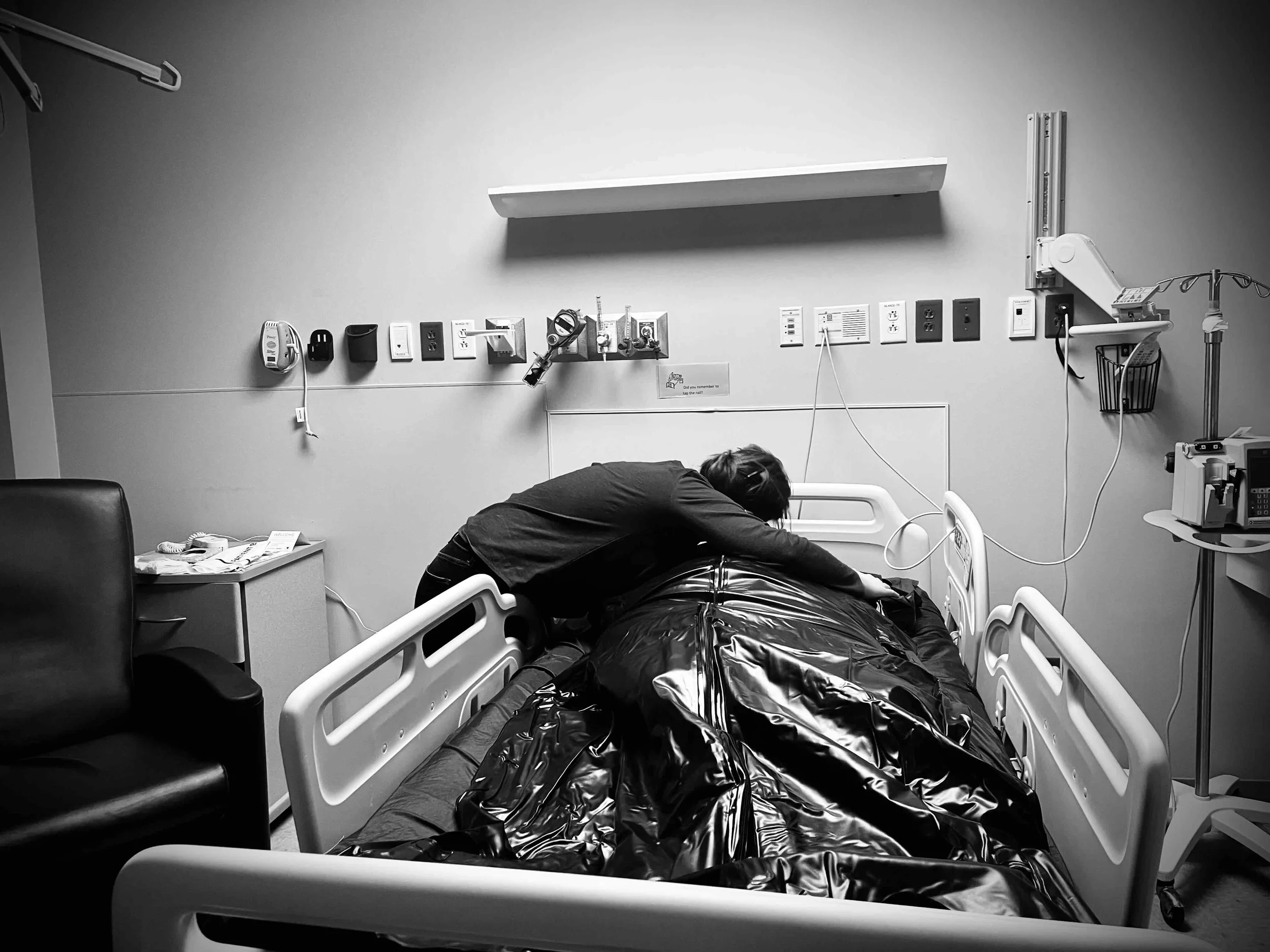 A person leaning over a hospital bed in a hospital room with medical equipment and a chair nearby.
