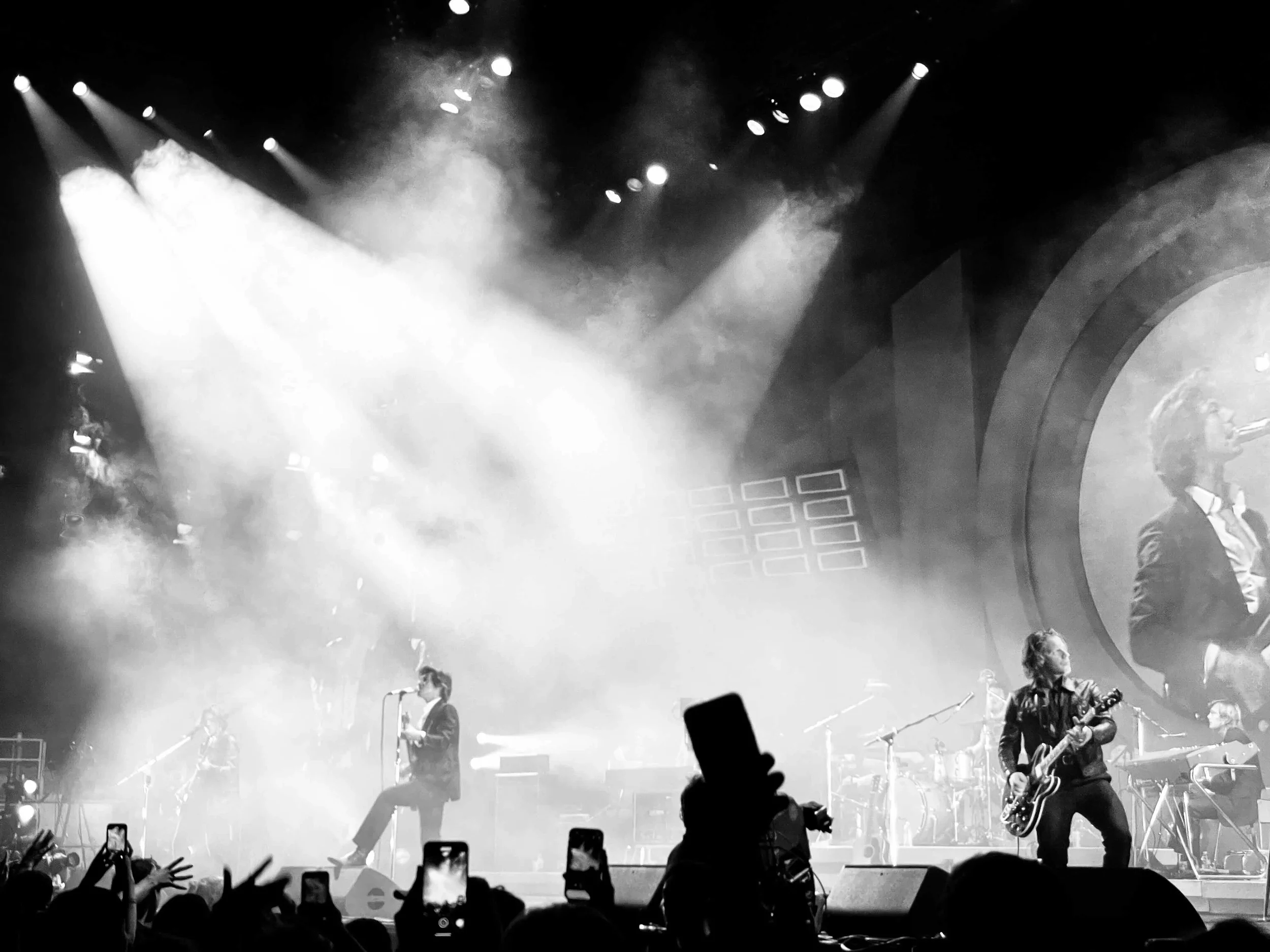 Black and white photo of a live music concert with a band performing on stage, crowd holding up phones, and stage lighting.