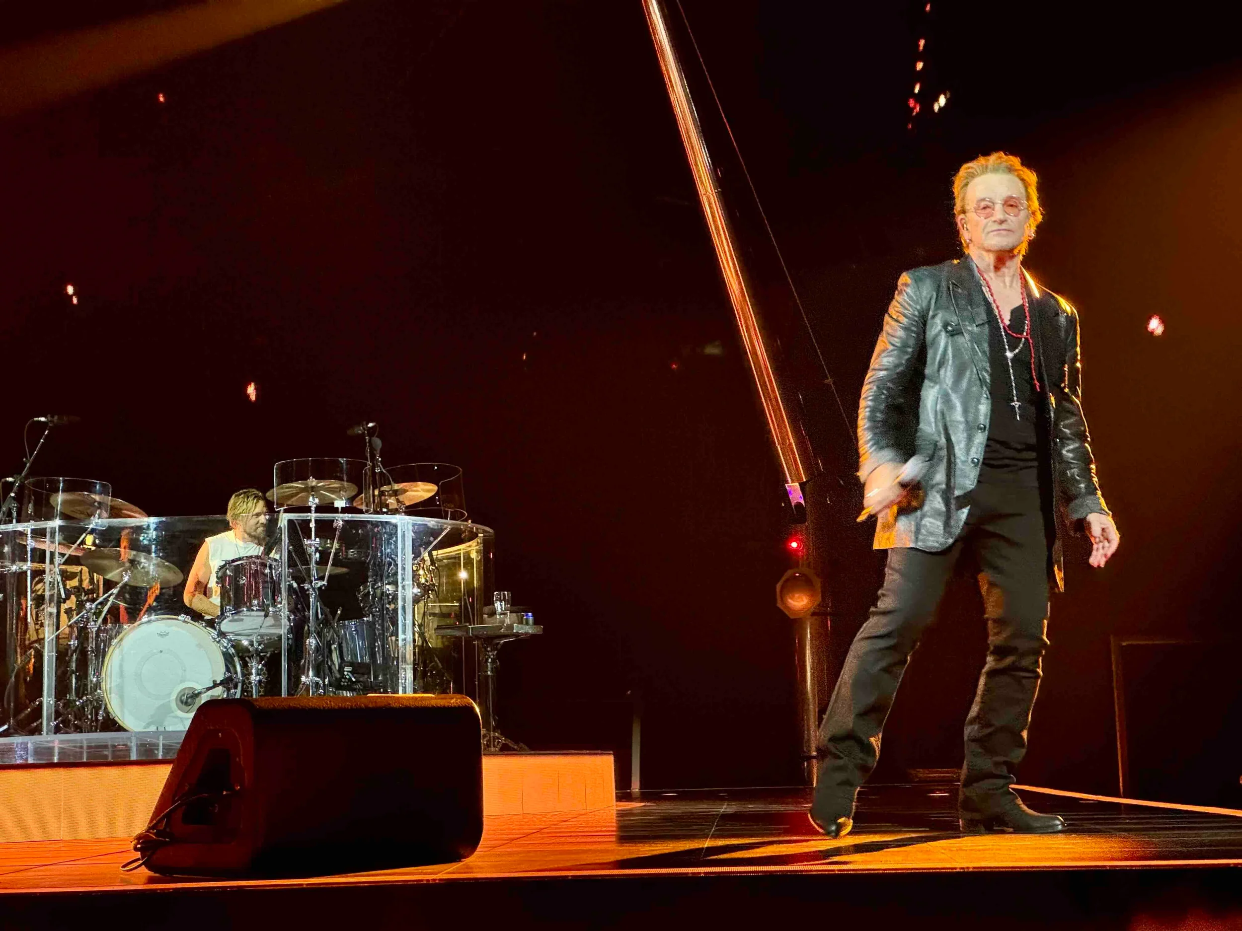 A rock singer with sunglasses, curly hair, a leather jacket, black shirt, black pants, and jewelry, standing on stage next to a drummer behind a clear drum shield, with stage lights and equipment visible.