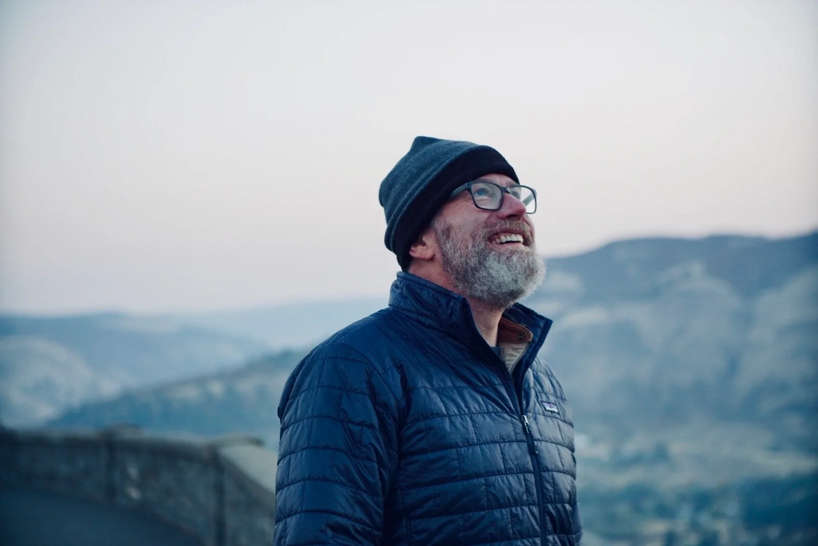 A bearded man wearing glasses, a blue puffy jacket, a black and gray knit beanie, smiling and looking upward outdoors with mountains in the background.