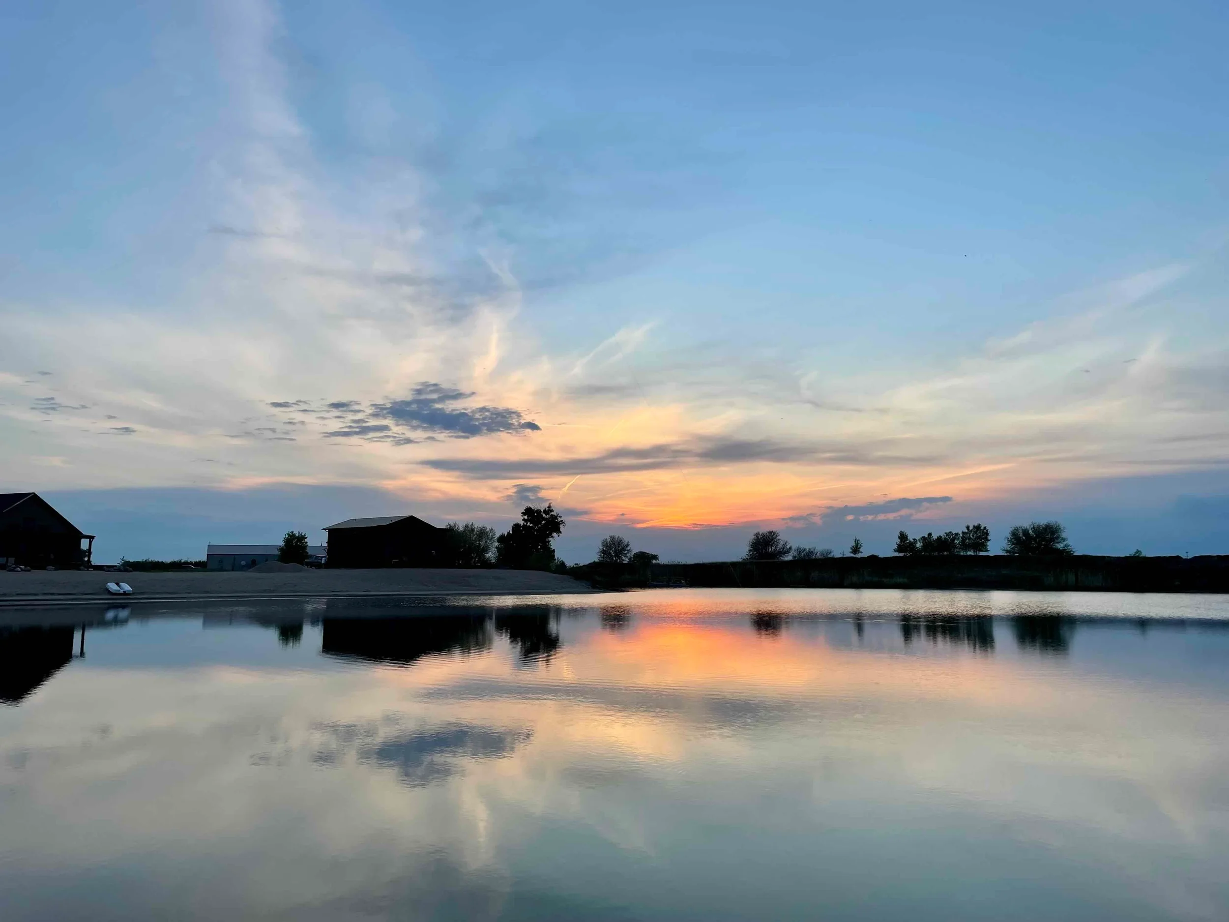 Serene sunset over a calm lake with reflections of the sky, trees, and barns