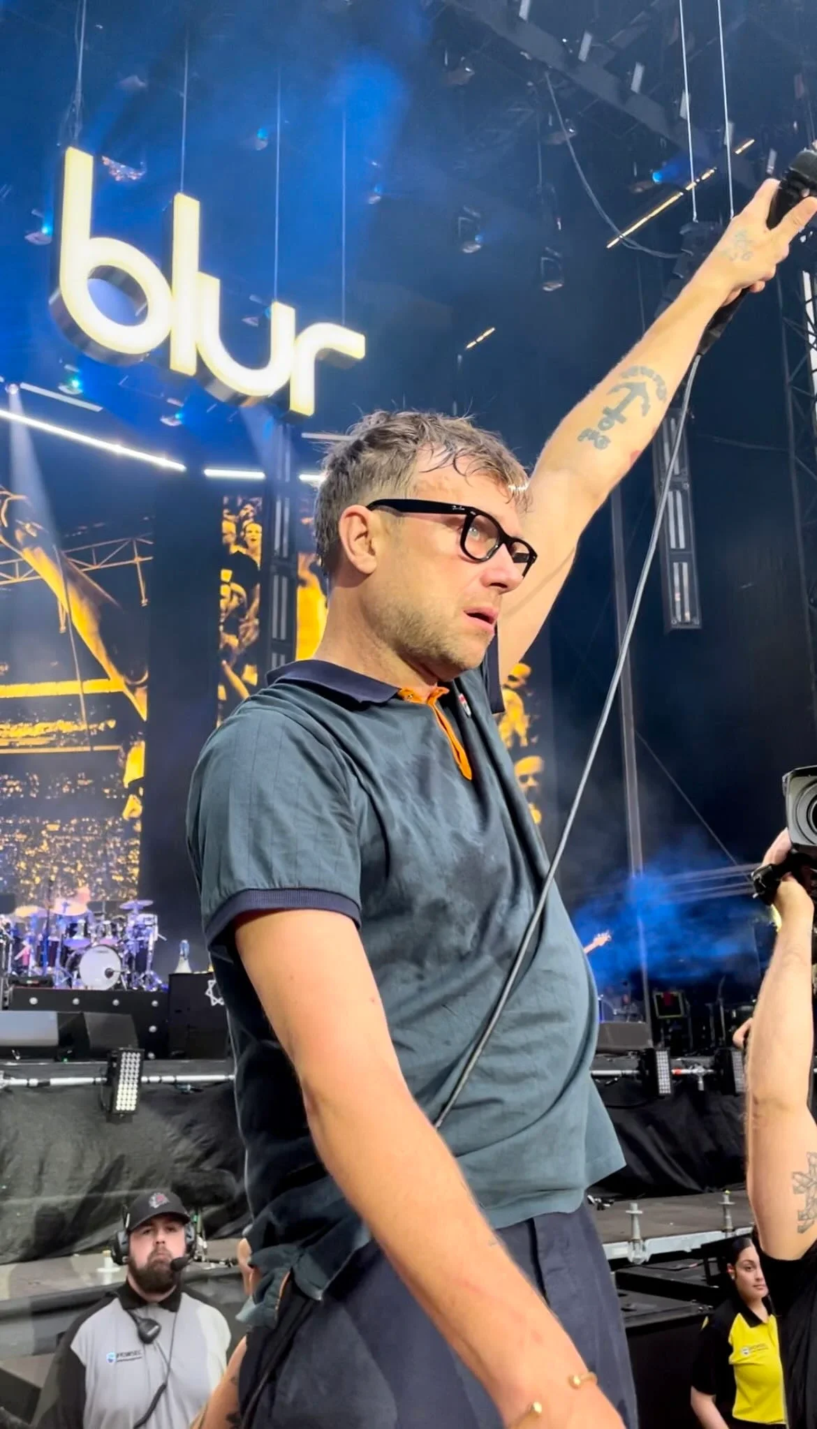 A man with glasses and tattoos on his arm is on stage at a concert, raising one arm in the air. The backdrop displays a large illuminated sign that reads 'blur' in lowercase letters.
