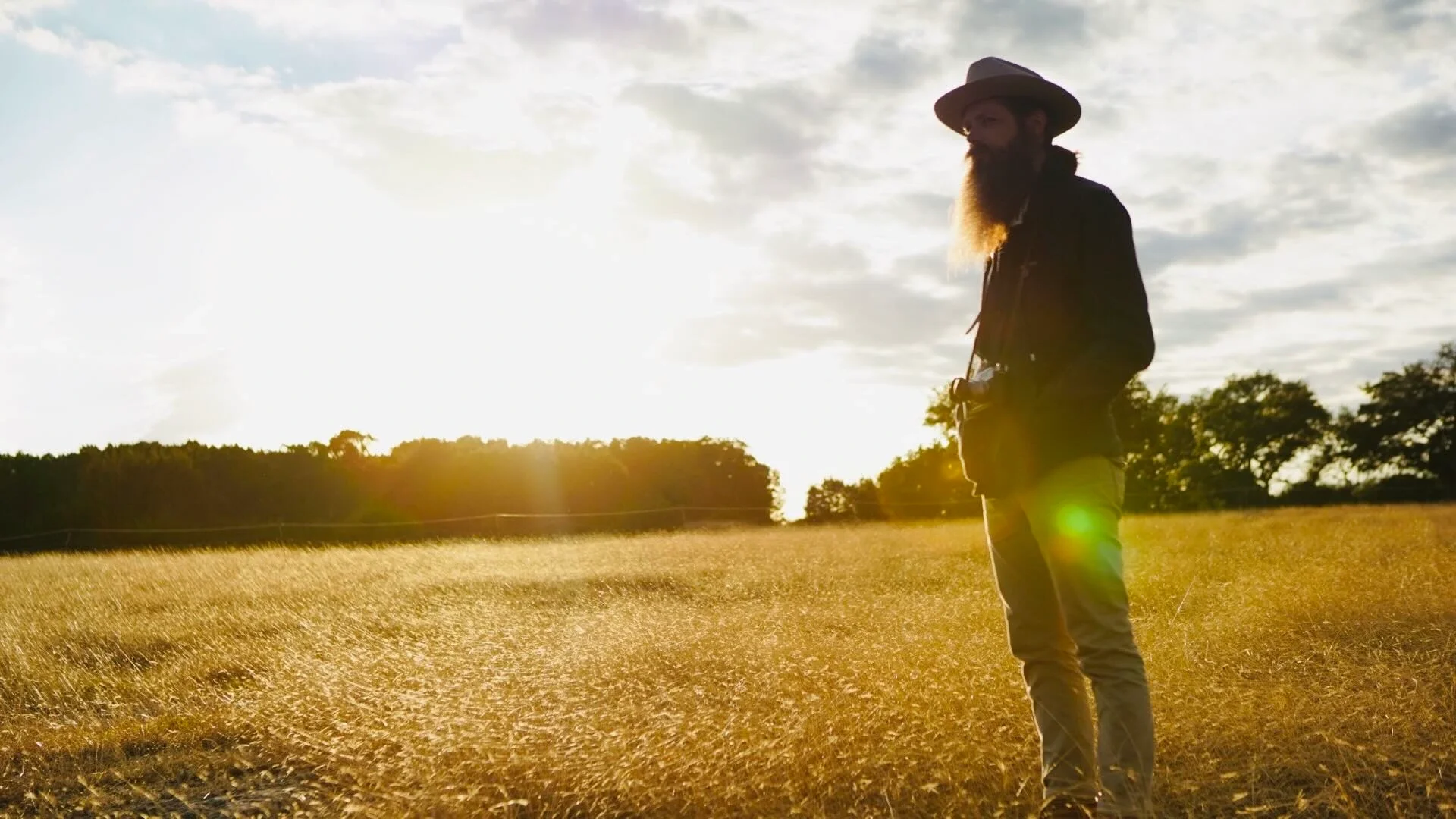 A man with a beard wearing a hat, dark jacket, and beige pants standing in a golden field during sunset, with trees in the background.