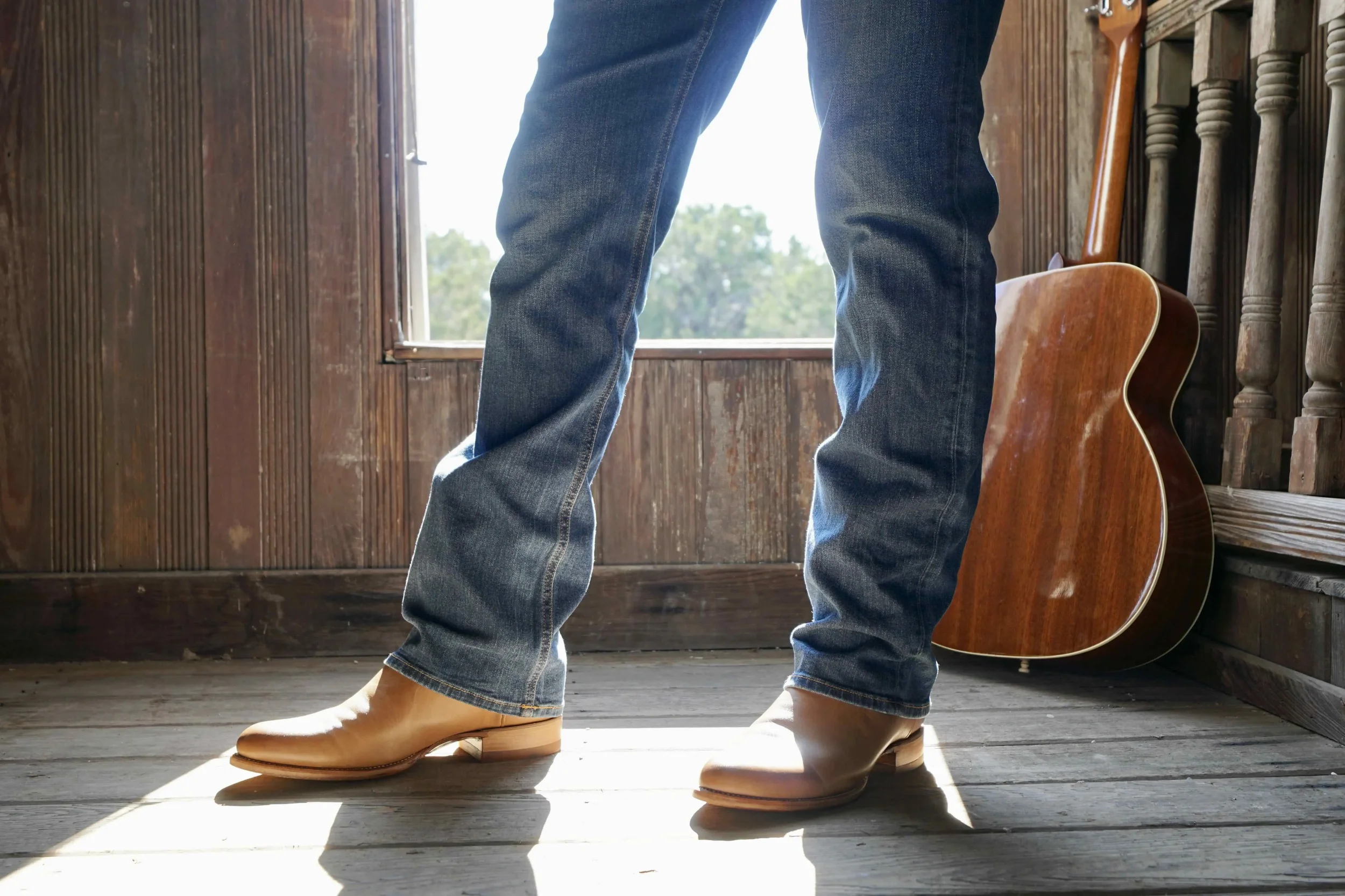 Person standing near a window wearing jeans and tan cowboy boots, with an acoustic guitar leaning against the wall in a wooden room.
