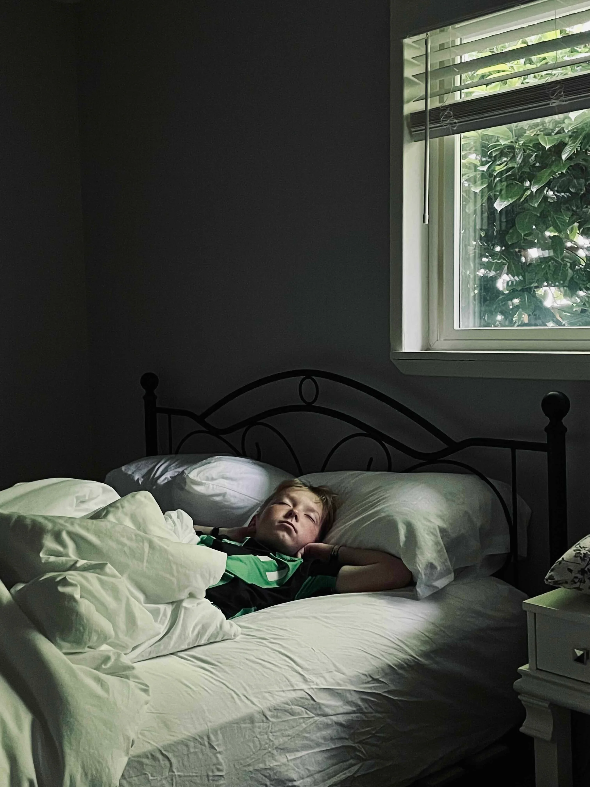 A boy sleeping in a bed with white sheets and pillows, by a window with blinds, showing greenery outside.
