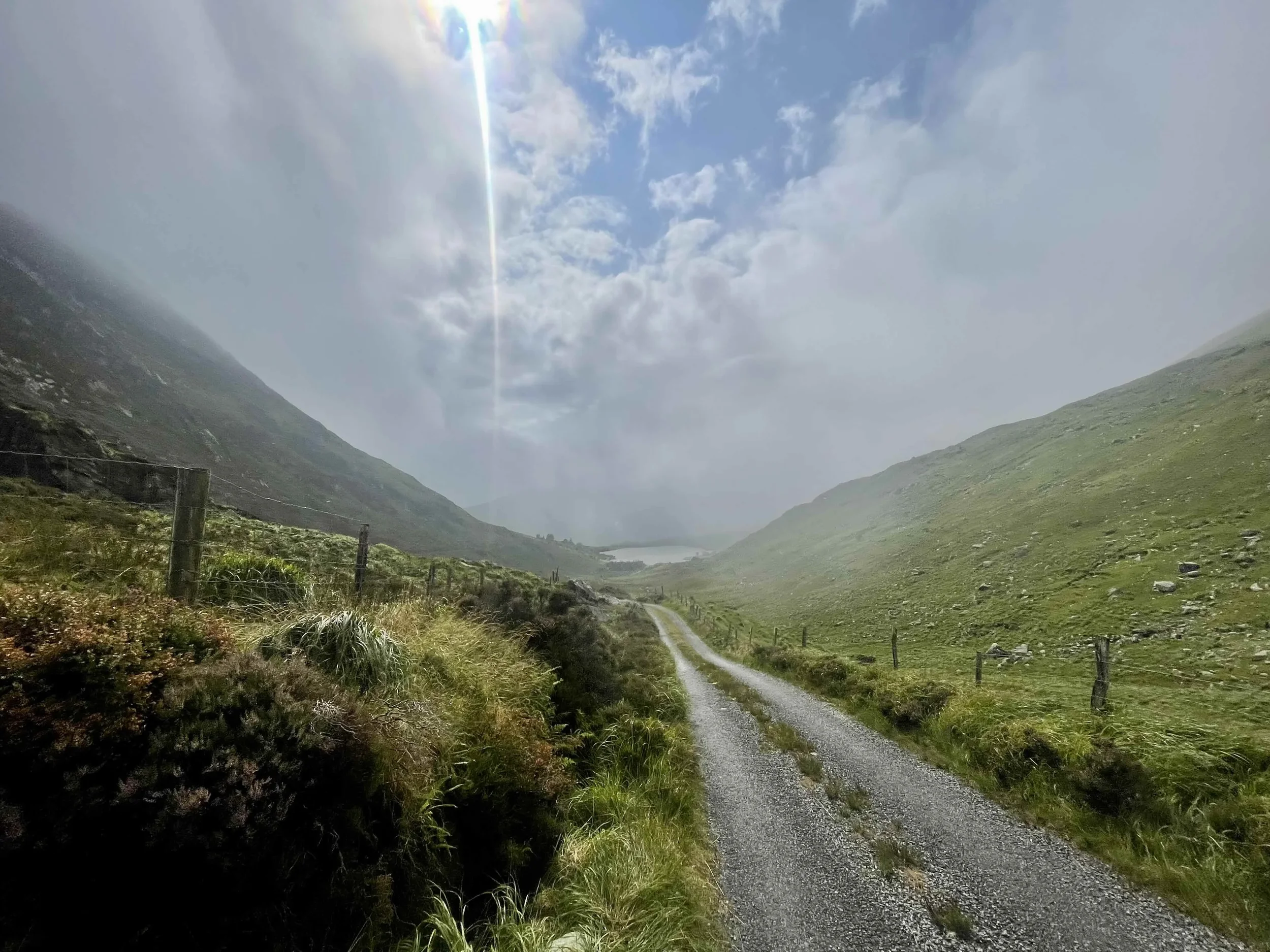 A gravel road winds through a green valley with grass and shrubs on both sides. Mountains with patches of snow rise on either side, and a small lake is visible in the distance. The sky is partly cloudy with sunlight streaming through a crack in the c