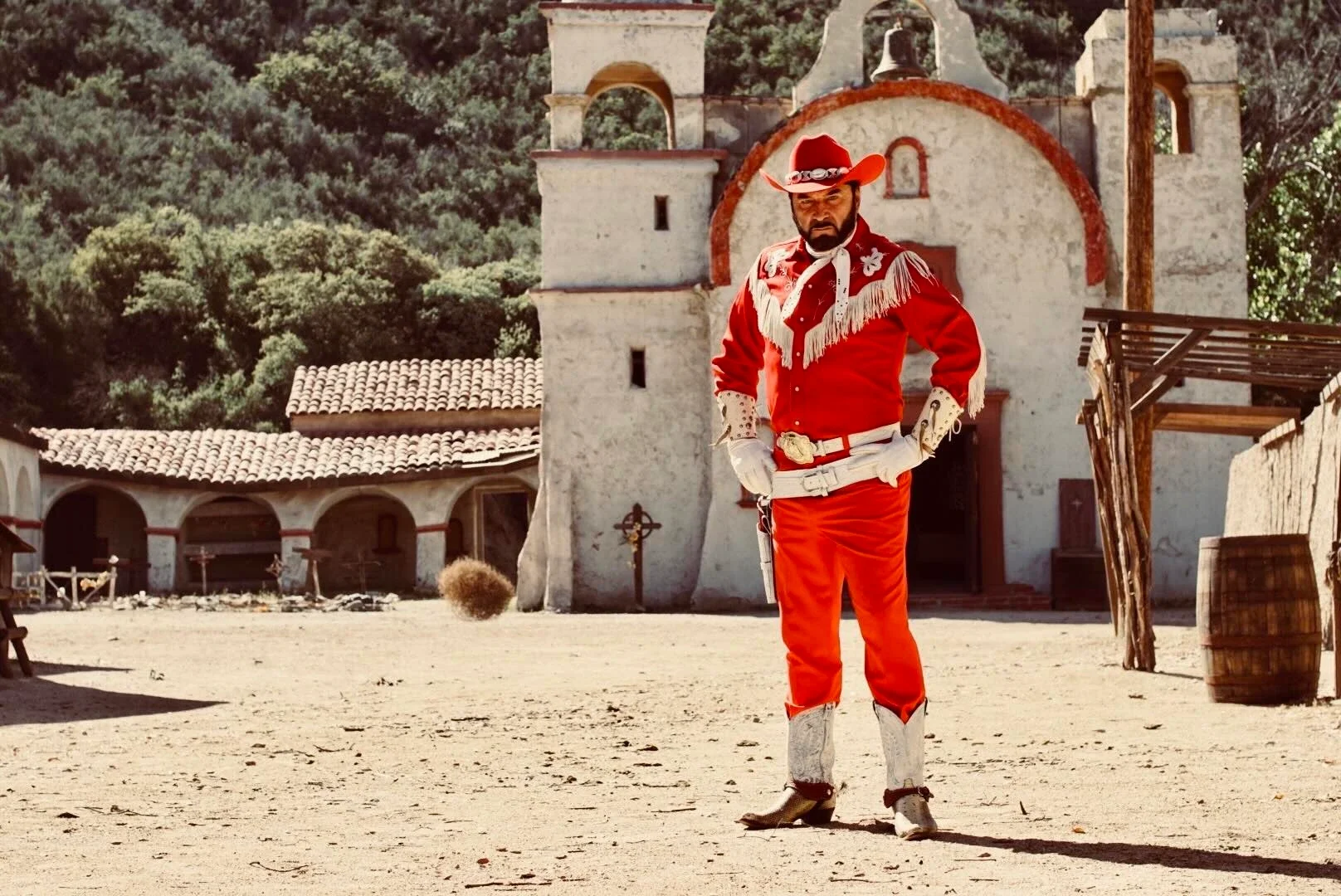 A man dressed as a traditional Mexican cowboy, standing outdoors in front of a historic mission-style church, wearing a red outfit with a wide-brimmed hat, decorated with white fringe and silver accents, with a serious expression and hands on hips.