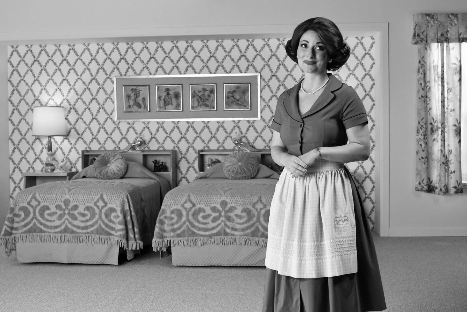 A woman dressed in vintage clothing standing in a bedroom with two beds, a bedside table with a lamp, and framed art on the wall.
