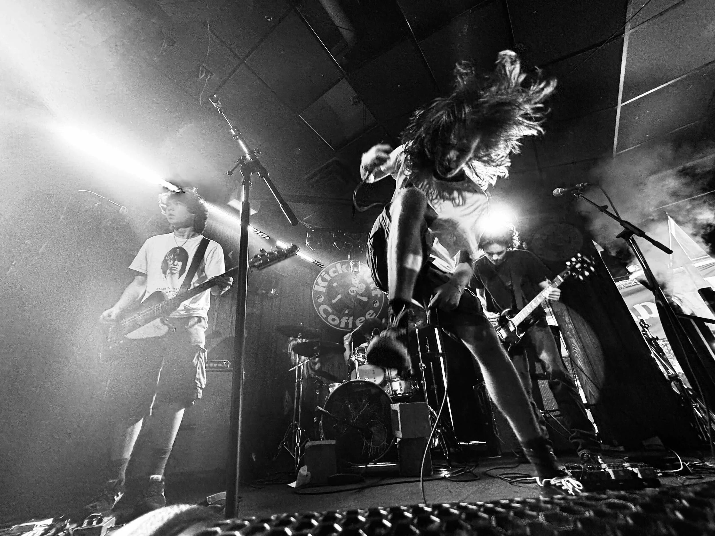A black-and-white photo of a live band performing on stage with energetic, dynamic movement. The lead singer is captured mid-jump with hair flying, holding a microphone. Two guitarists are playing, one on the left with a bass guitar and one on the ri