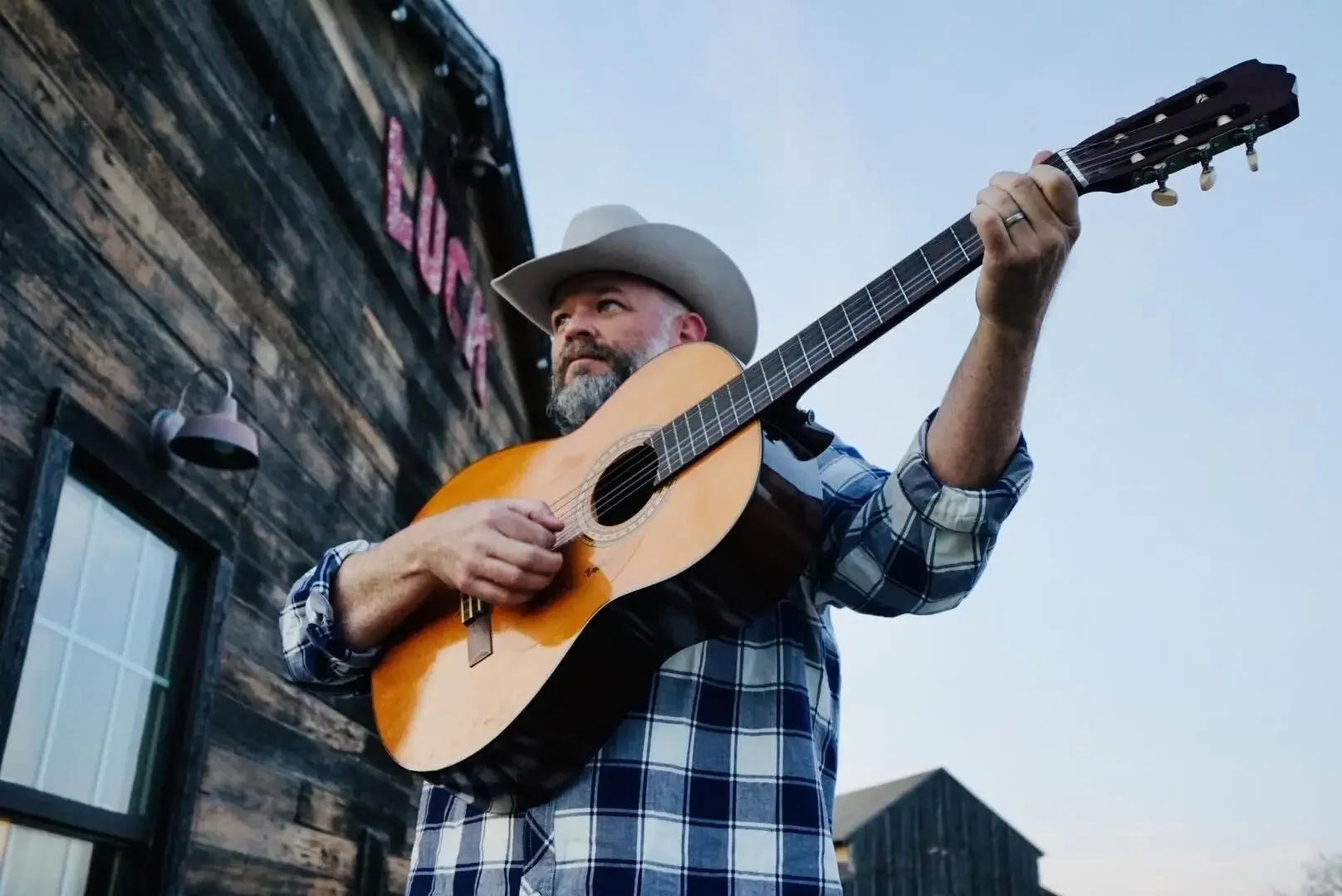 A man wearing a cowboy hat and plaid shirt playing an acoustic guitar outdoors during the daytime.