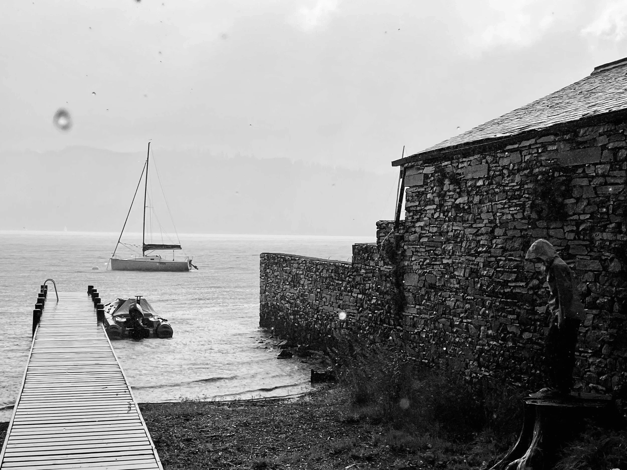 A black and white photo of a boat docked at a pier, with a stone building on the right. A person is standing on a bench next to the building, looking down. The water is calm with a sailboat visible in the distance, and there is a mountain range or fo
