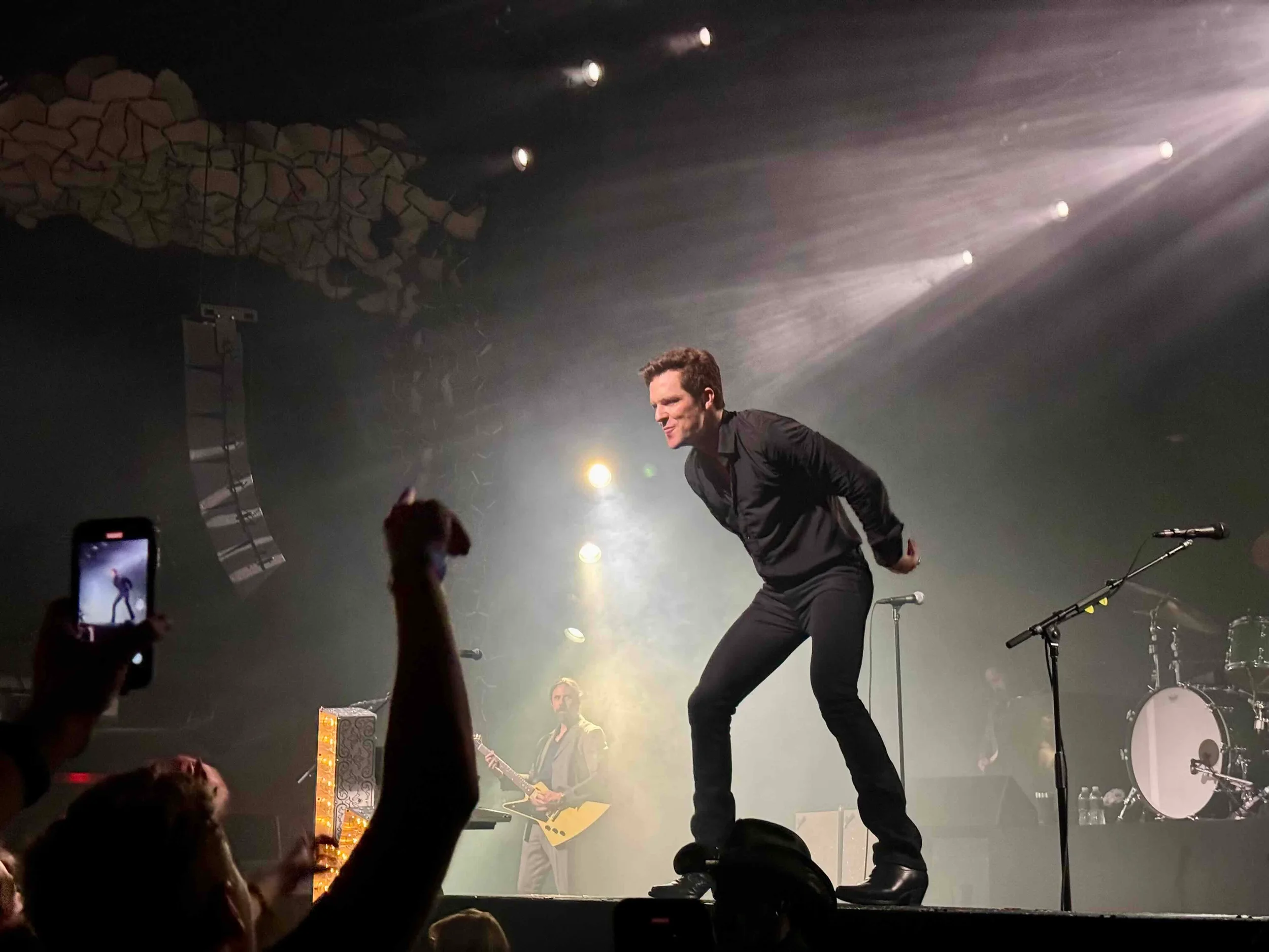 A singer performing on stage with a microphone stand, with a guitarist and drums in the background, and an audience taking photos.