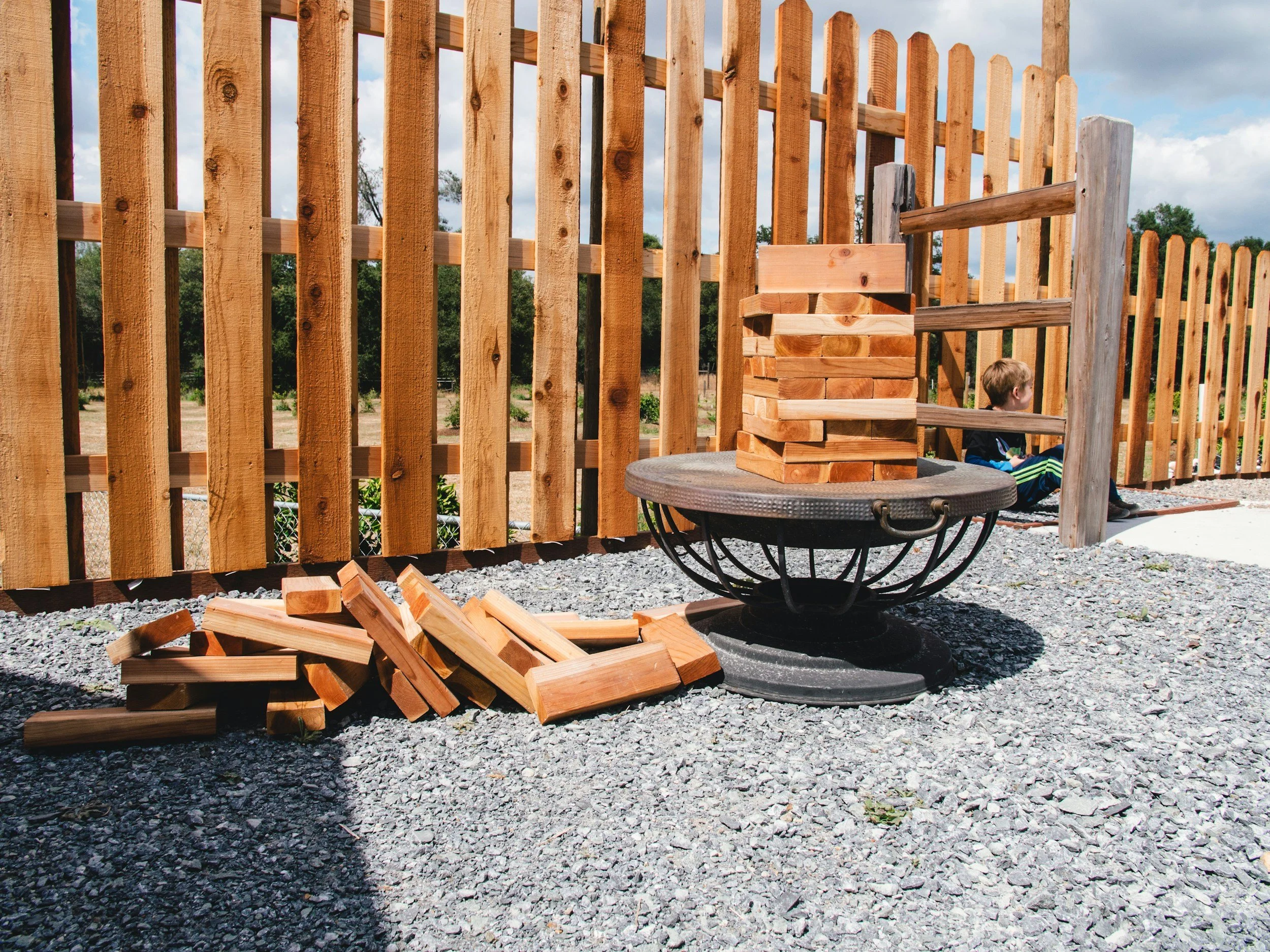 A child sitting on the ground near a wooden fence with a pile of wooden planks and a decorative fire pit with a stack of wood on top.