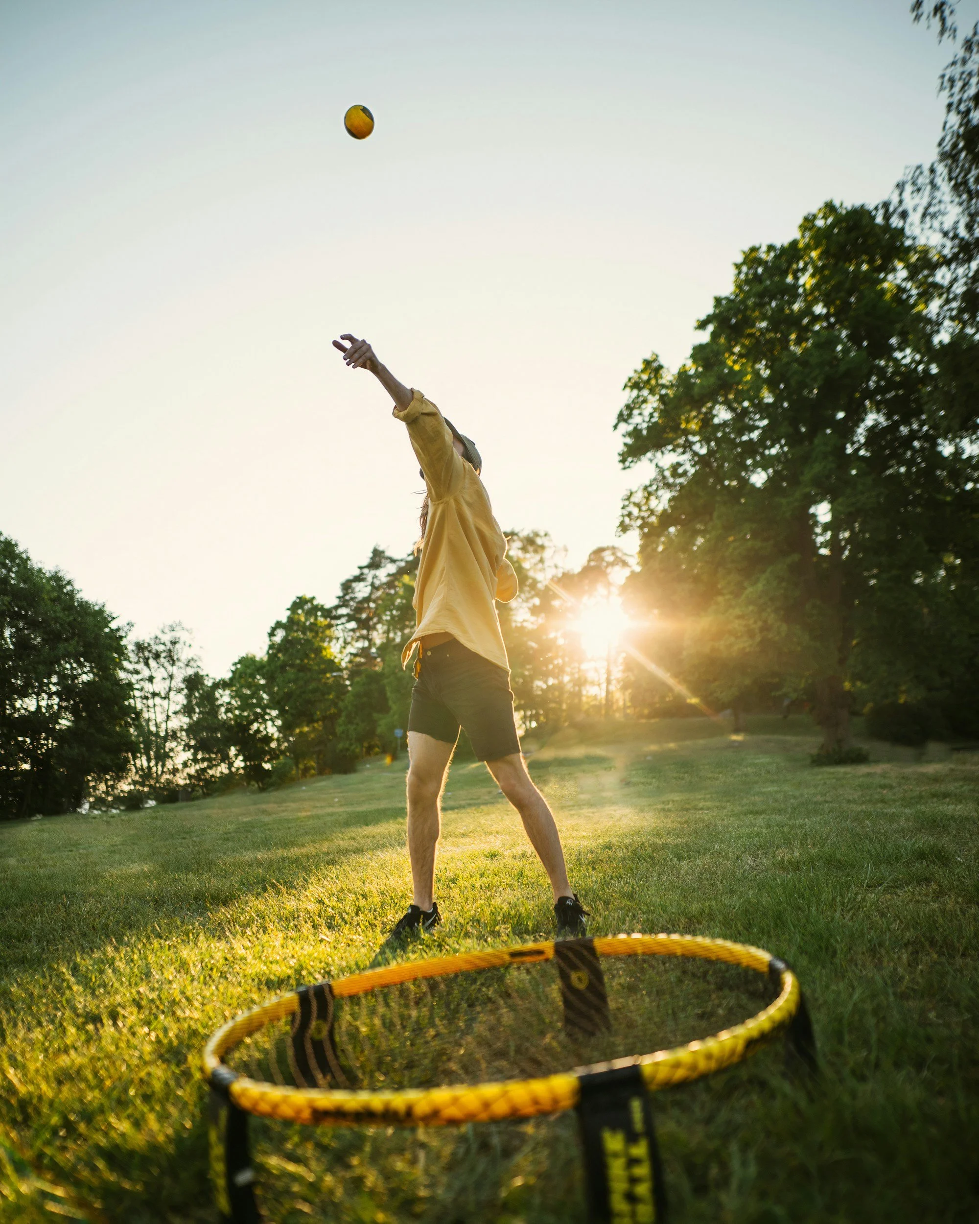 A person playing a game of catch with a yellow ball outdoors during sunset, with a yellow and black frisbee on the grass in the foreground, surrounded by trees and grass.