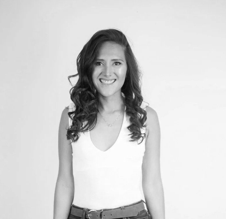 Jovenc woman with long, curly hair, sonriendo, usando una camiseta blanca y jeans, en un fondo simple.