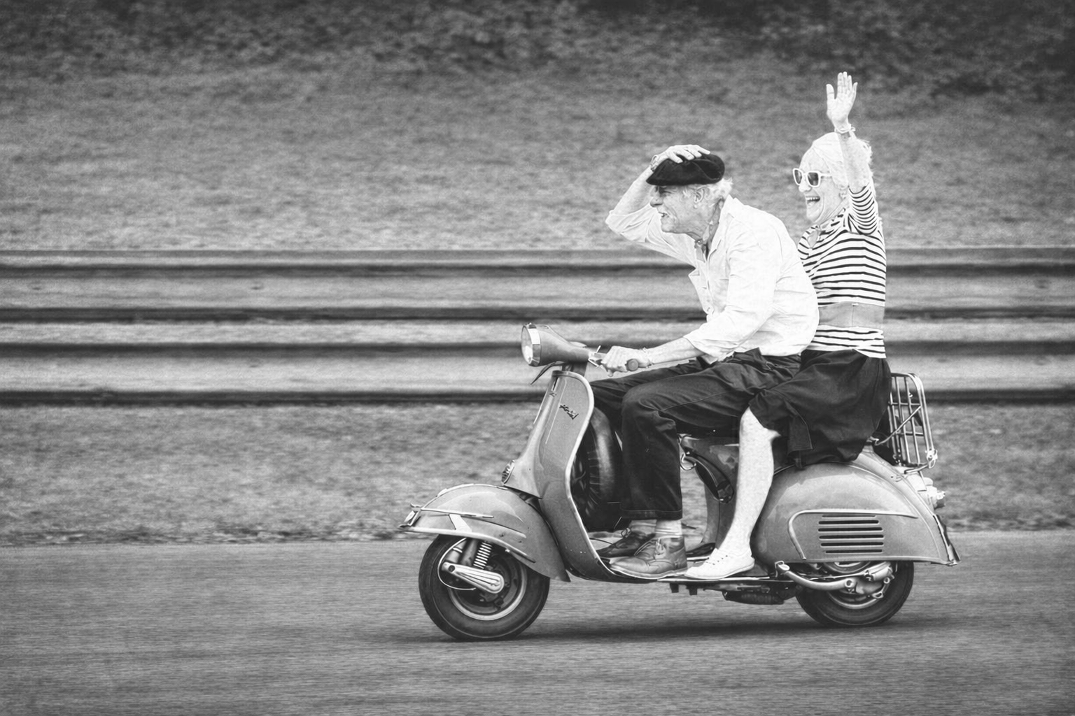 Two elderly people, a man and a woman, riding a vintage scooter together on a road near water, smiling and waving, in black and white.