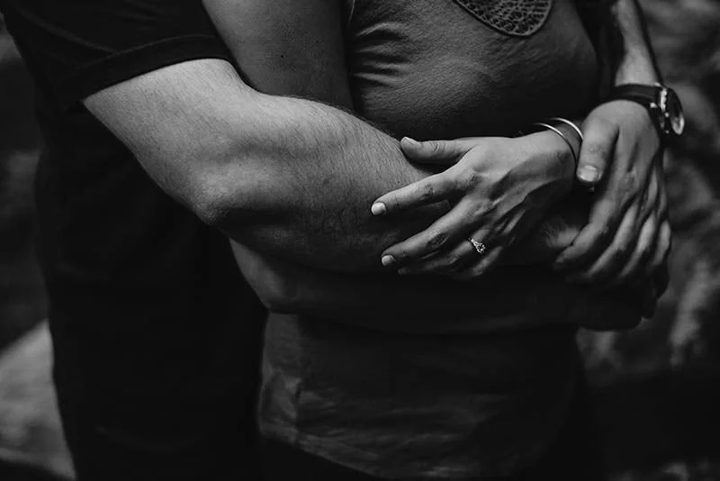 Close-up of a couple embracing, with the man's arm around the woman's waist and the woman's hands resting on his arm, in black and white.