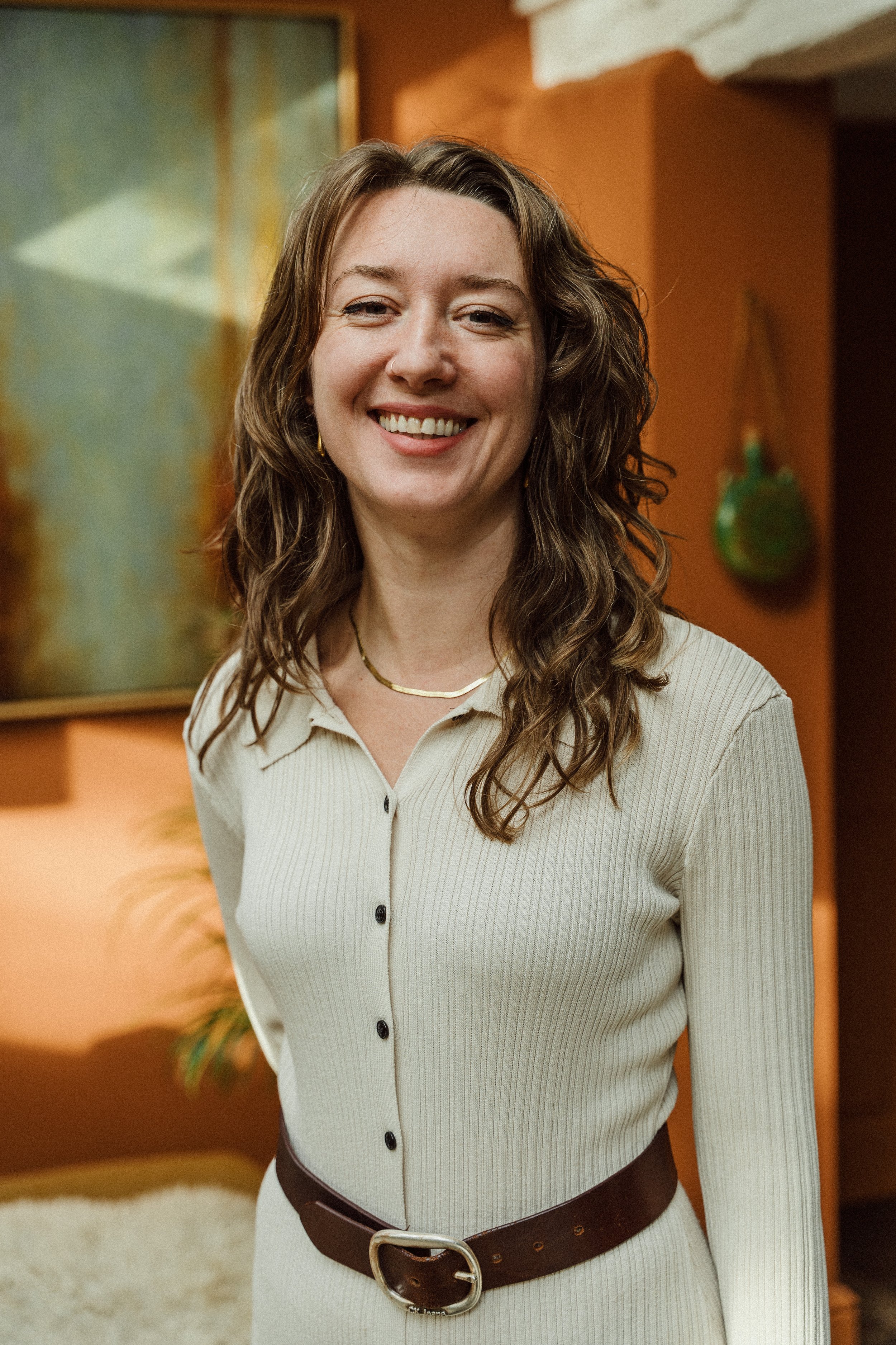 A woman with wavy brown hair smiling, wearing a cream-colored button-up dress with a dark brown belt, standing indoors with a painting on the wall behind her.