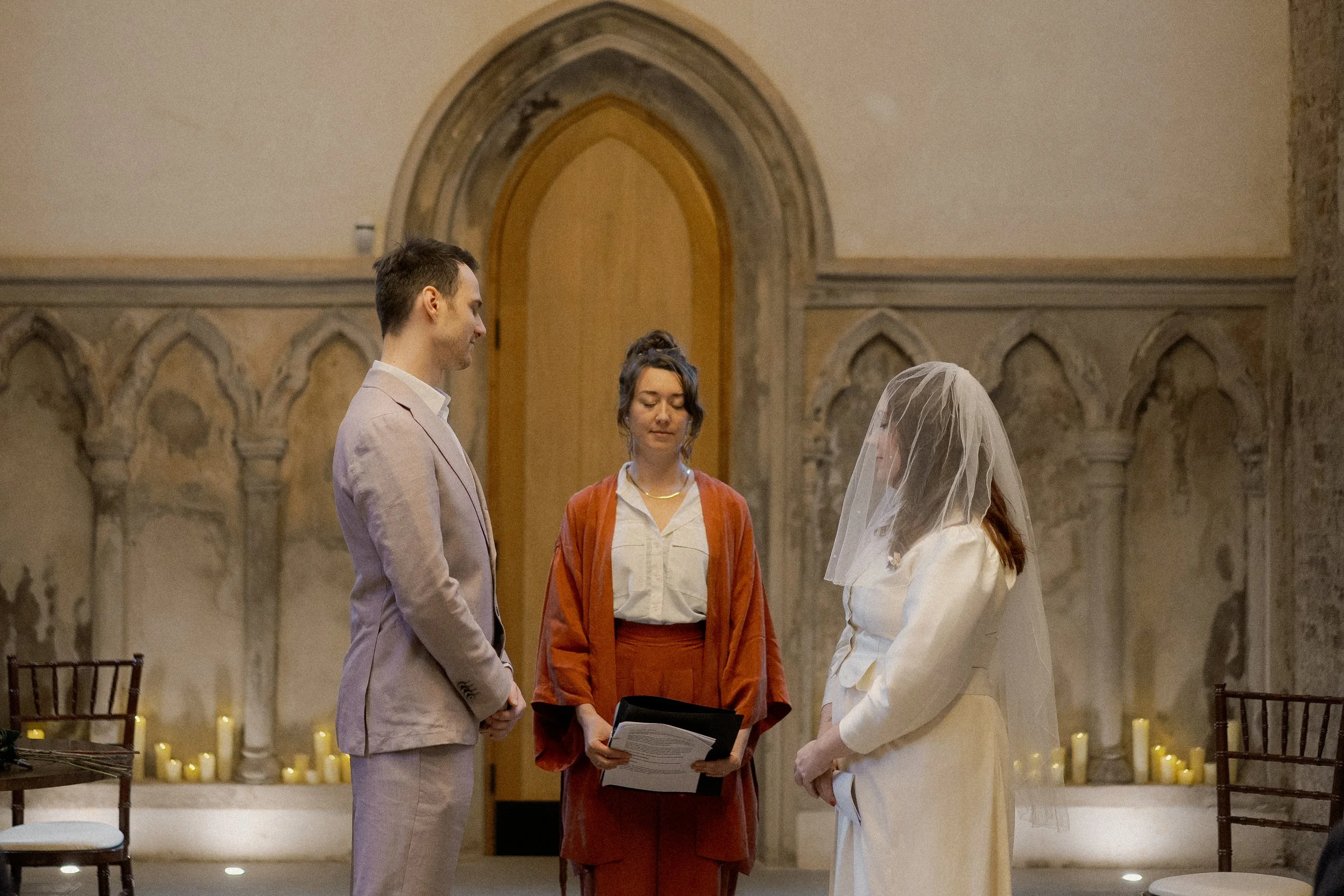 A wedding ceremony with a bride and groom standing opposite each other in a church, with an officiant holding a book, inside a historic stone building decorated with candles.