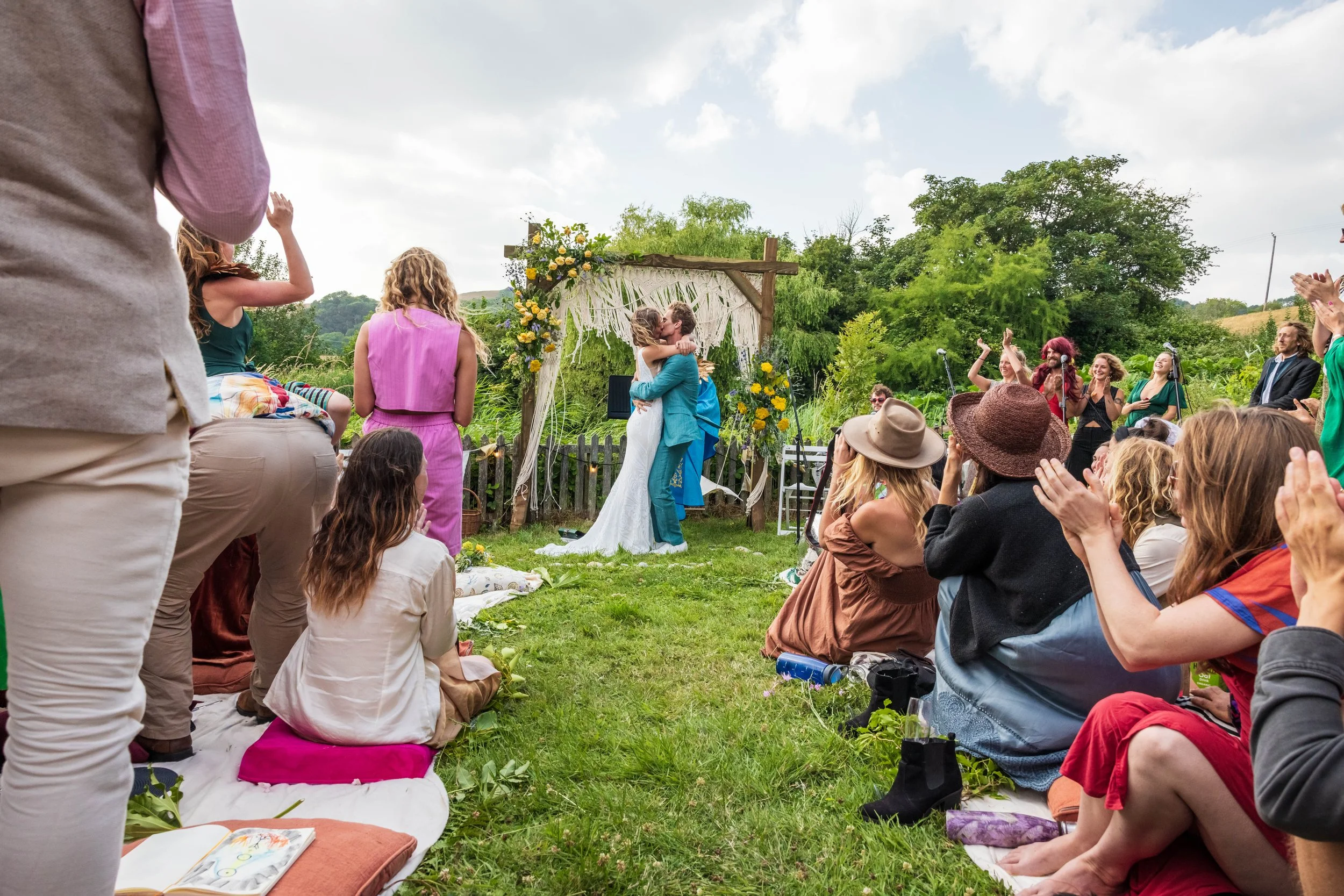 A wedding ceremony taking place outdoors with a bride and groom kissing under a decorated archway, surrounded by seated guests and greenery.