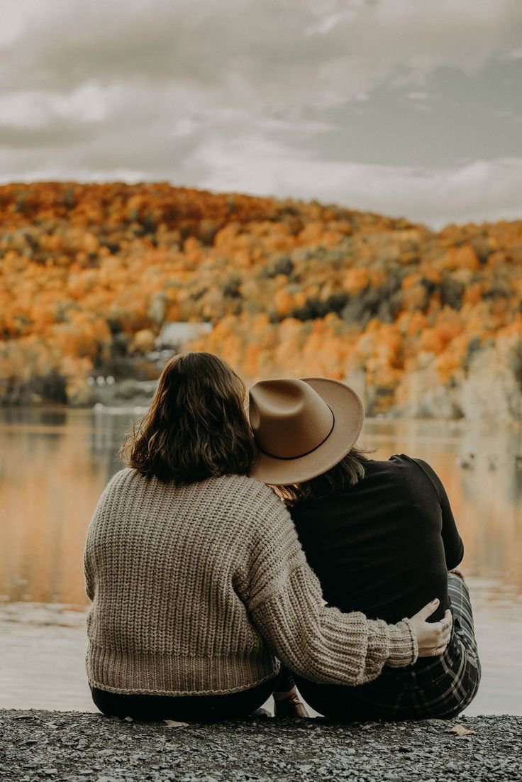 Two women sitting by a lake during autumn, one wearing a gray sweater and the other a hat and black shirt, with colorful trees in the background.
