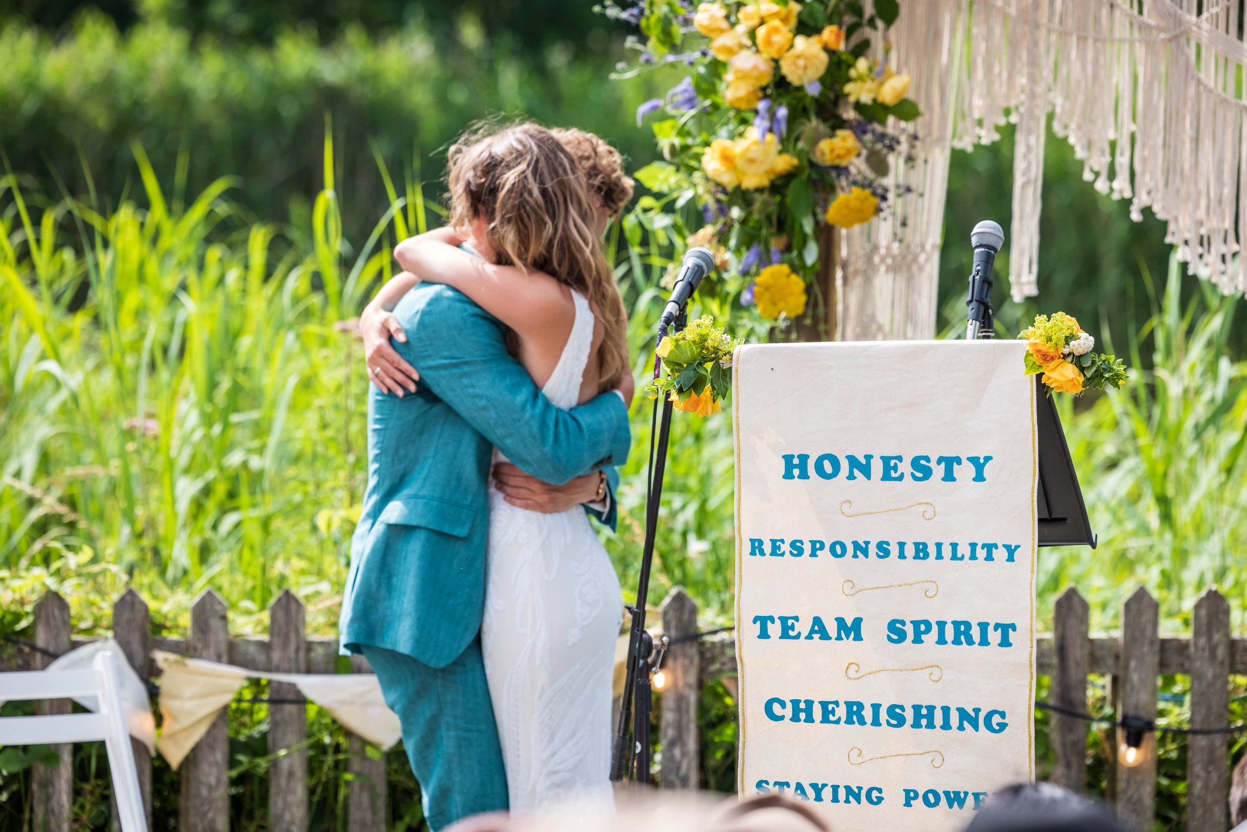 A couple hugging at an outdoor wedding ceremony surrounded by greenery, with a wedding sign displaying words like honesty, responsibility, team spirit, and cherishing.