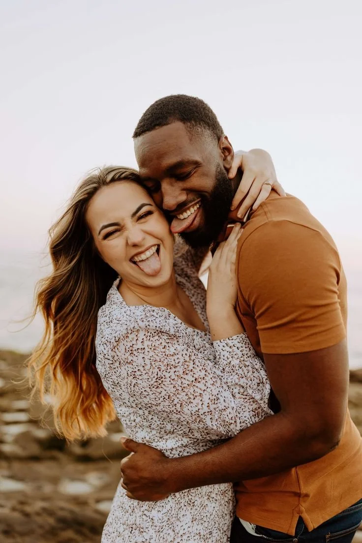 A happy couple hugging and making silly faces outdoors, with the woman sticking out her tongue and the man smiling with his tongue out, during daytime.