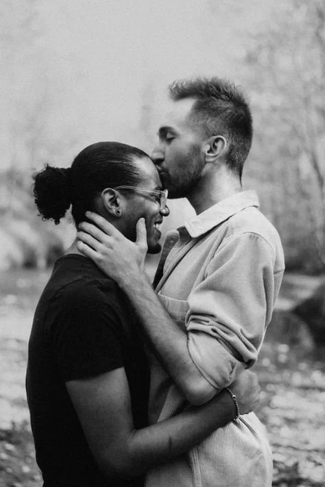 A couple sharing a tender moment outdoors, with the man kissing the woman's forehead while she smiles and holds his arms.