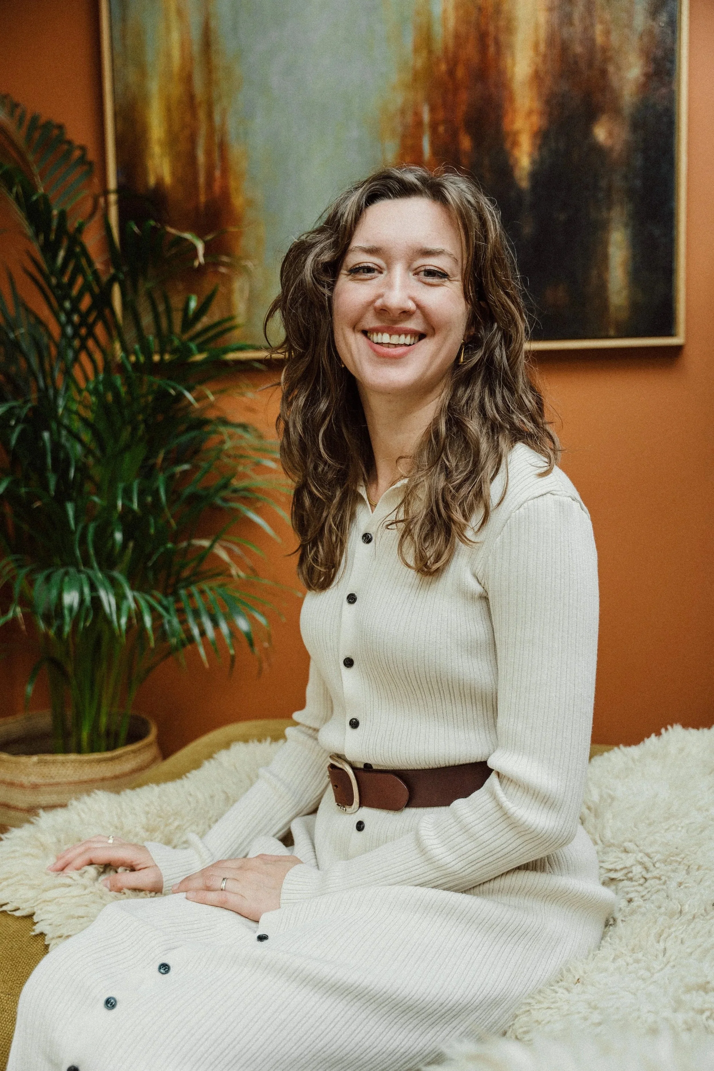 A woman sitting on a cozy beige blanket with a smile, wearing a cream-colored ribbed dress with black buttons and a wide brown belt, in a room with a large green plant, a colorful abstract painting, and orange walls.