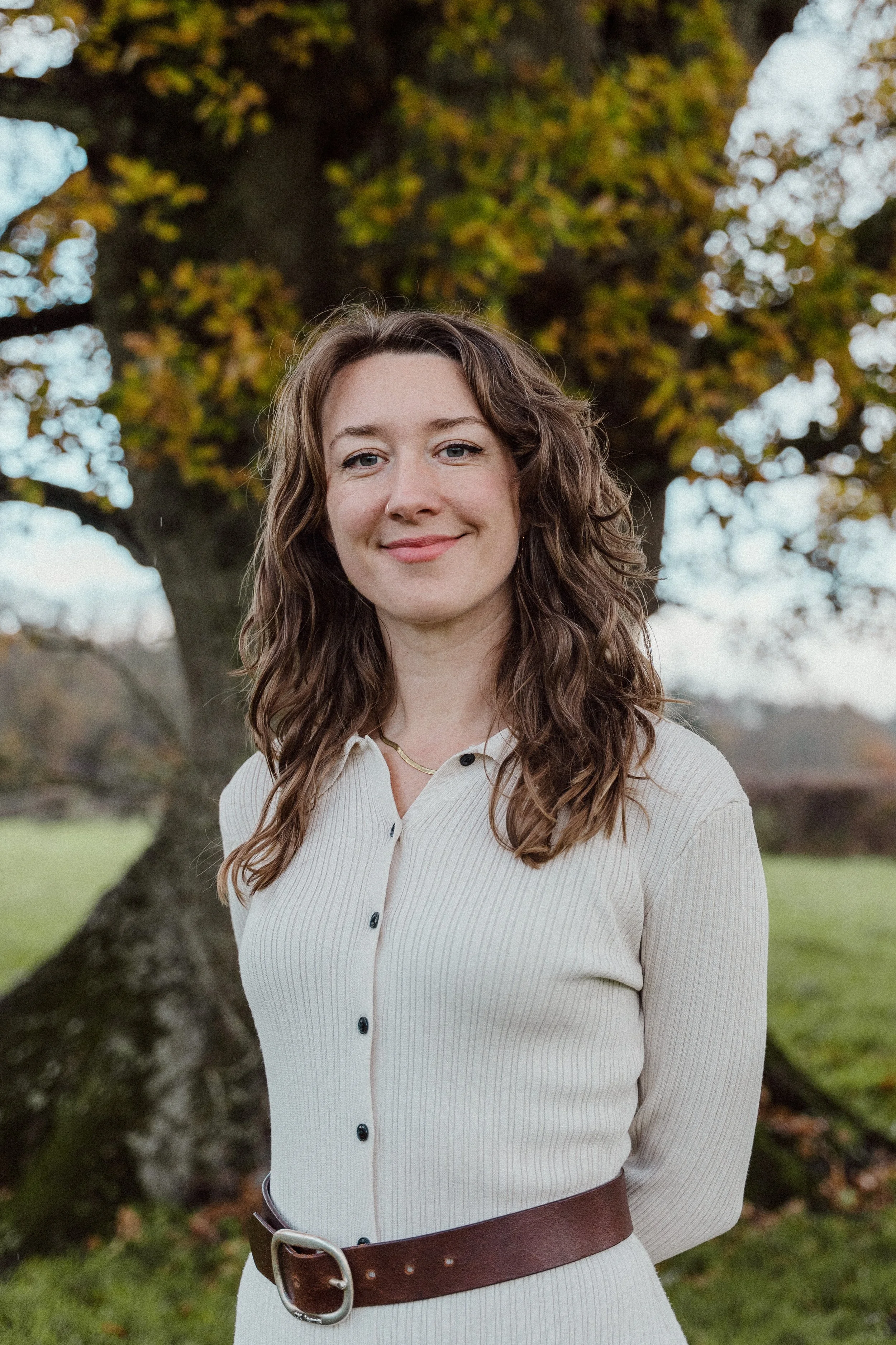 A woman with wavy brown hair standing outdoors near a tree with orange and green leaves, wearing a cream-colored ribbed long-sleeve shirt with black buttons and a brown belt, smiling at the camera.