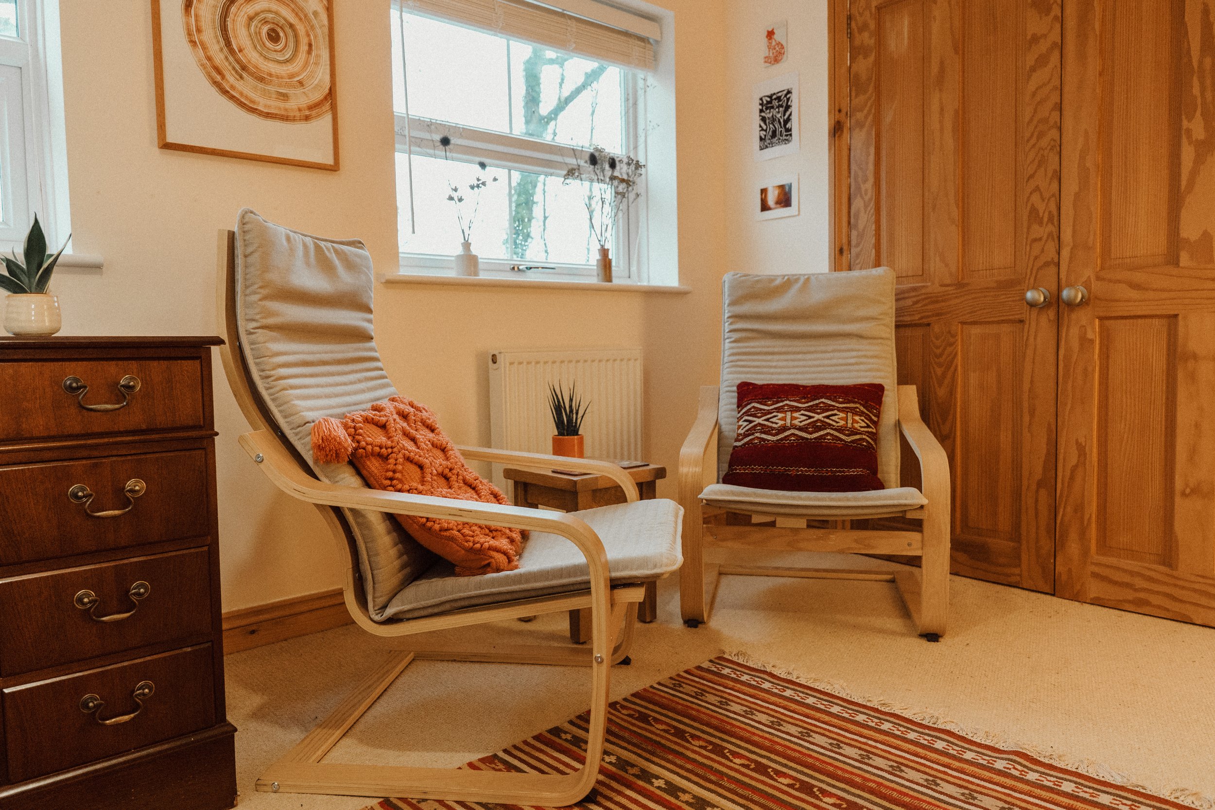A cozy corner with two beige armchairs with wooden frames, each having a decorative pillow, one orange and one with a patterned design. A small side table with a plant is between the chairs. To the left, part of a dark wooden dresser with drawers is visible, and on the wall above it, framed artwork hangs. There is a window with blinds, a plant on the windowsill, and a wooden closet with two doors on the right. A colorful striped rug is on the floor.