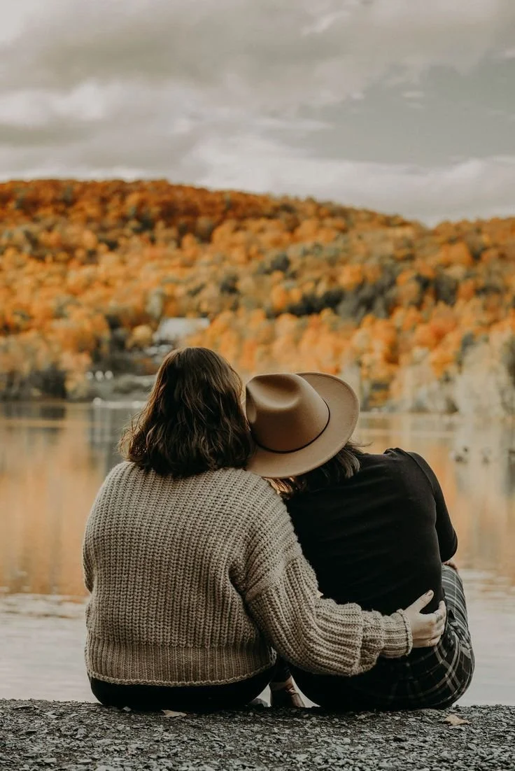 Two women sitting by a lake, one wearing a beige sweater and the other in a black shirt and plaid pants, with a scenic autumn landscape of trees with orange leaves in the background.