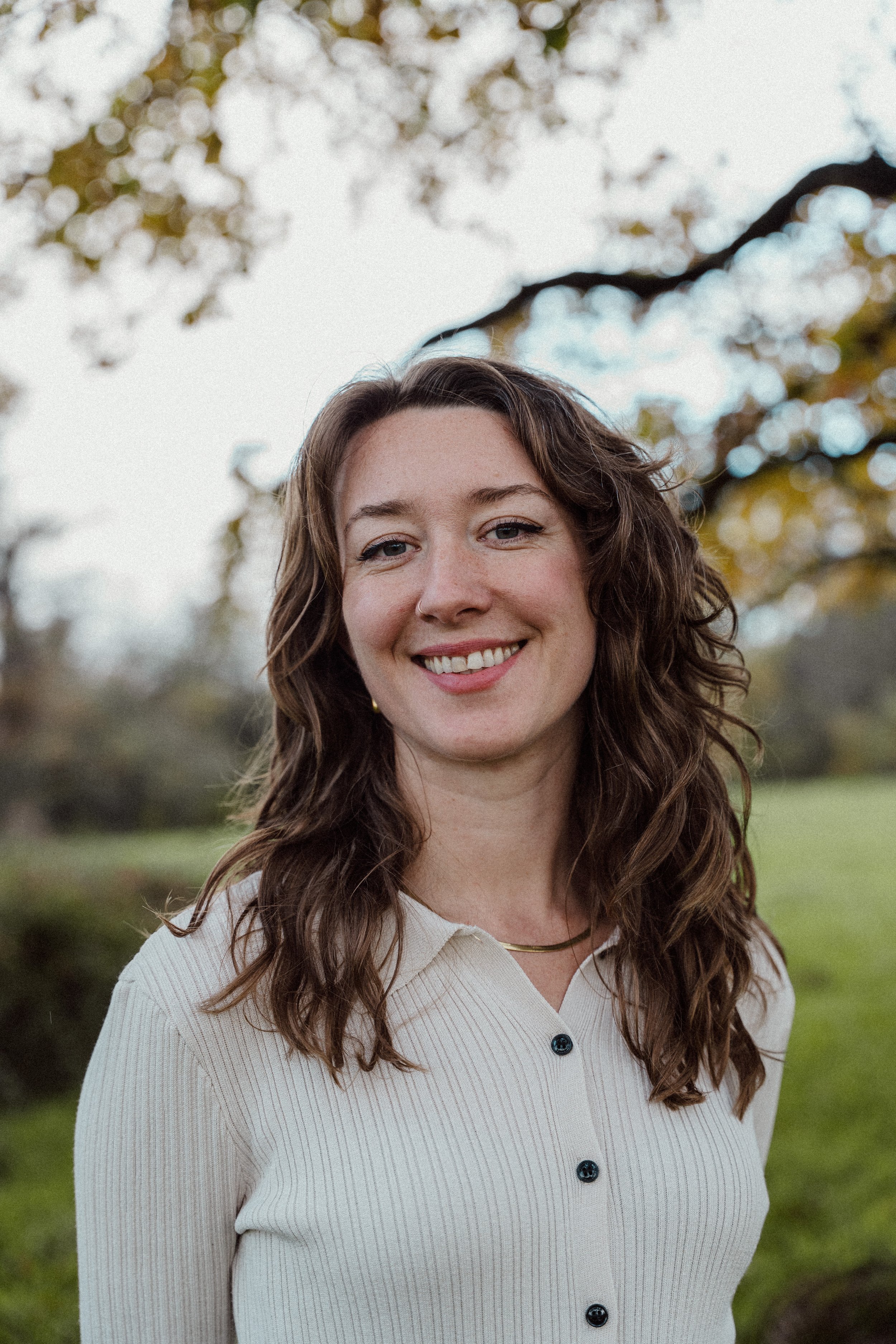 A young woman with wavy brown hair, smiling, standing outdoors with trees and a cloudy sky in the background.