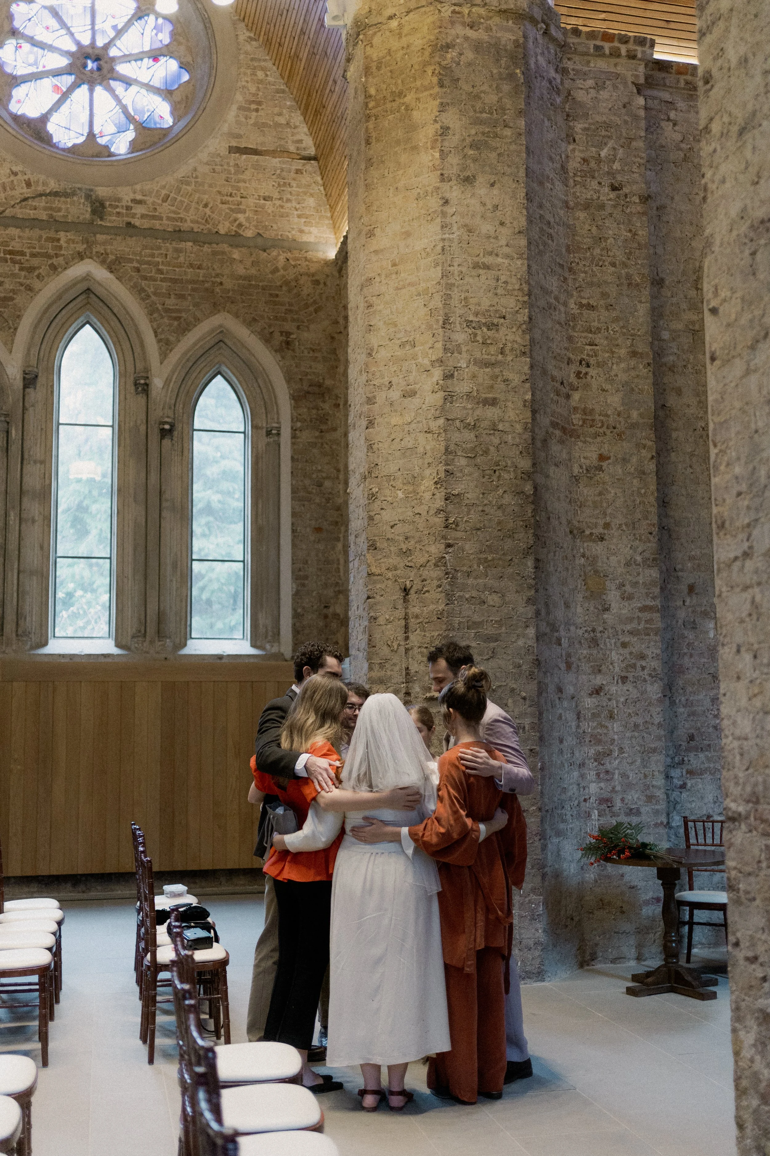 A group of people wearing formal attire hugging each other in a church with stained glass windows and brick walls.