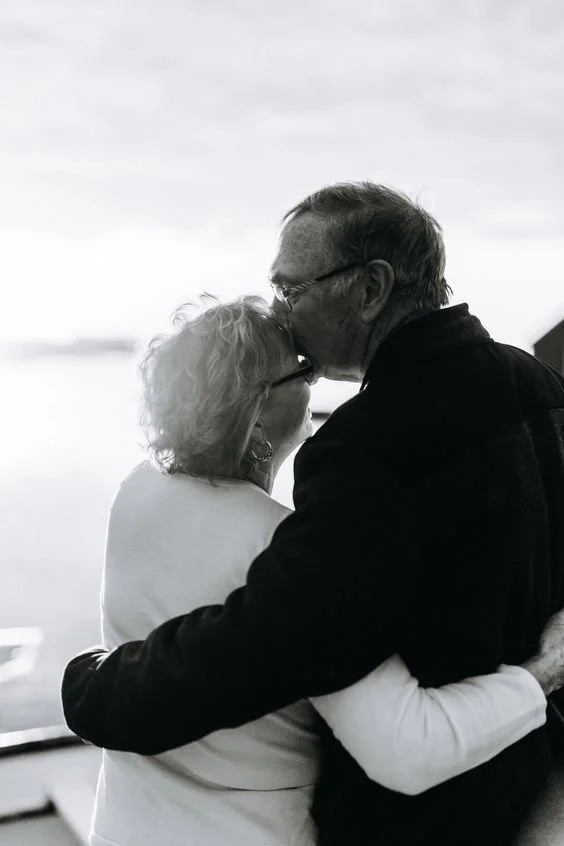 An elderly couple embracing and kissing each other indoors with a scenic window view in the background.