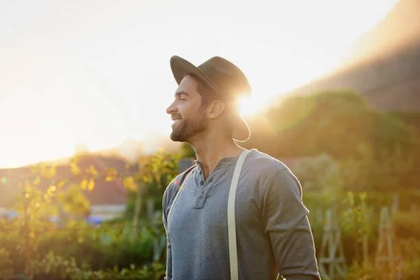 A man with a hat smiling outdoors during sunset, carrying a backpack in a rural setting.
