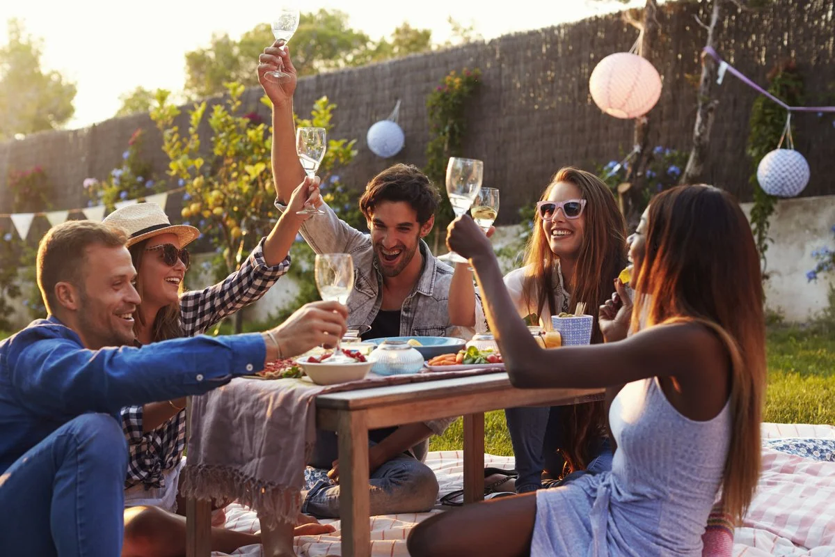 Group of friends enjoying a backyard picnic, raising glasses in a toast, sitting on a blanket with food and drinks, decorated with paper lanterns and bunting.