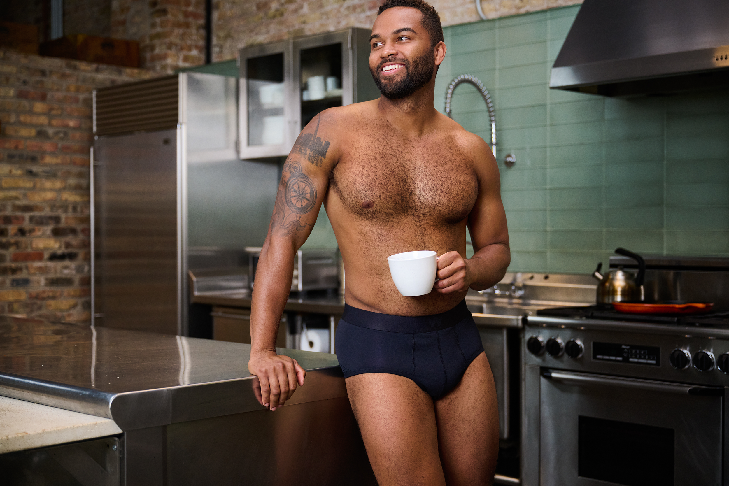 A man with a beard, tattoos on his arm and chest, wearing only underwear, standing in a modern kitchen holding a white mug and smiling.