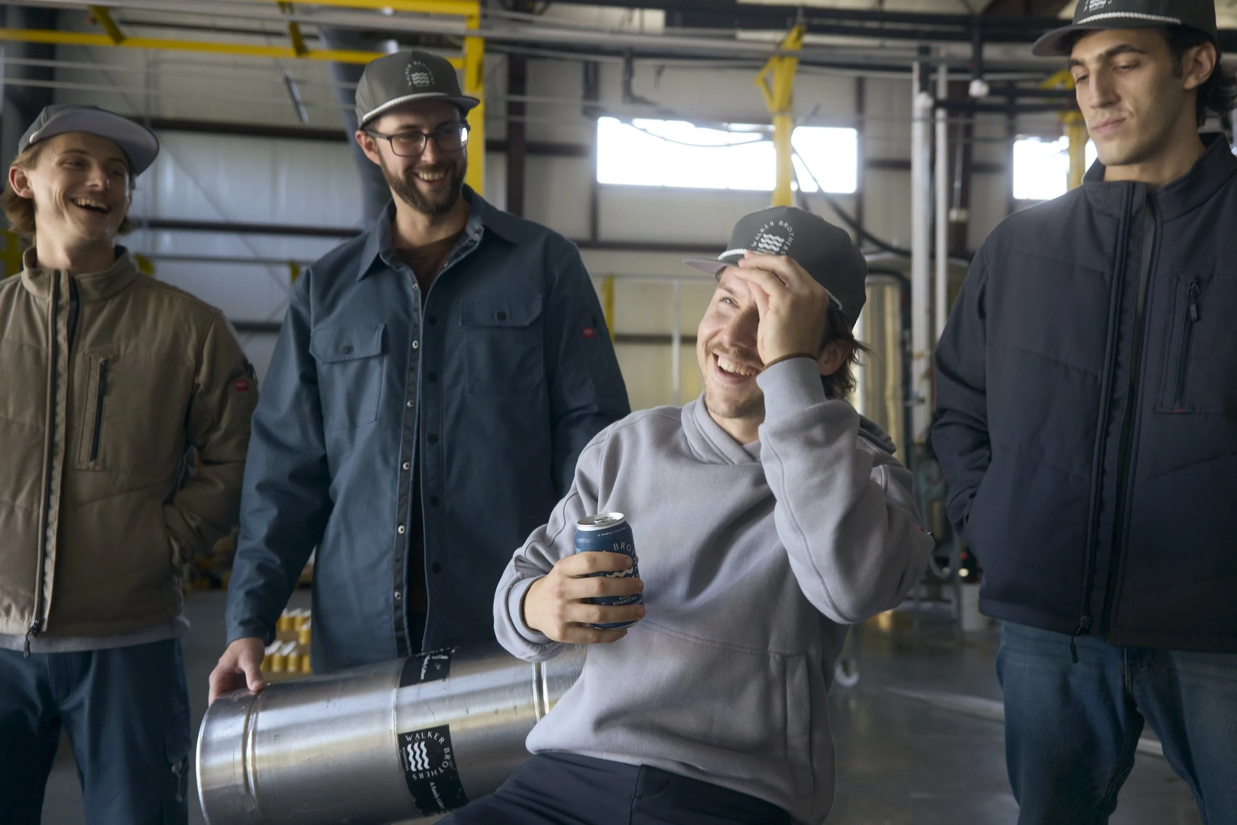 Group of five young men in casual clothing and baseball caps inside an industrial warehouse, smiling and laughing with one holding a metal pipe and a beverage can.