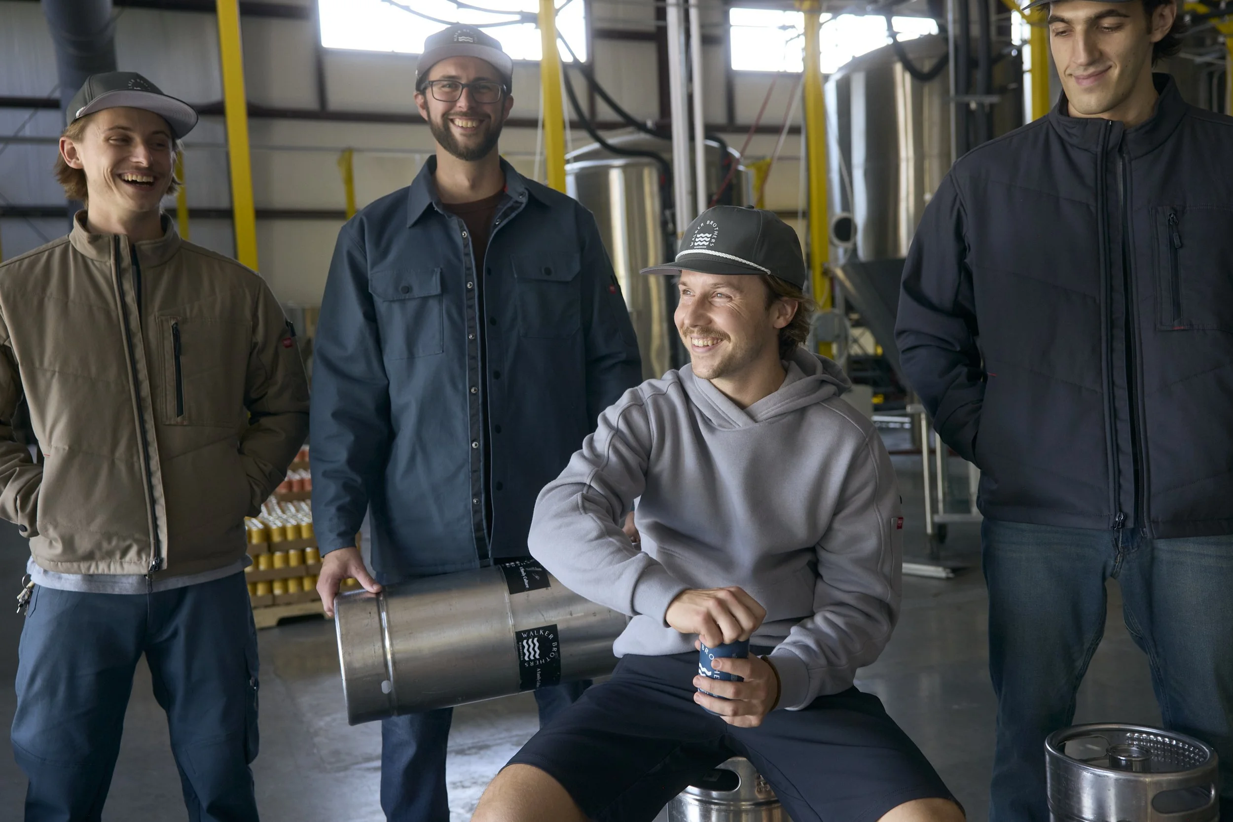 Four young men in casual clothing with jackets and caps inside an industrial or brewery setting, smiling and gathered around a large metal barrel, with one sitting and others standing.