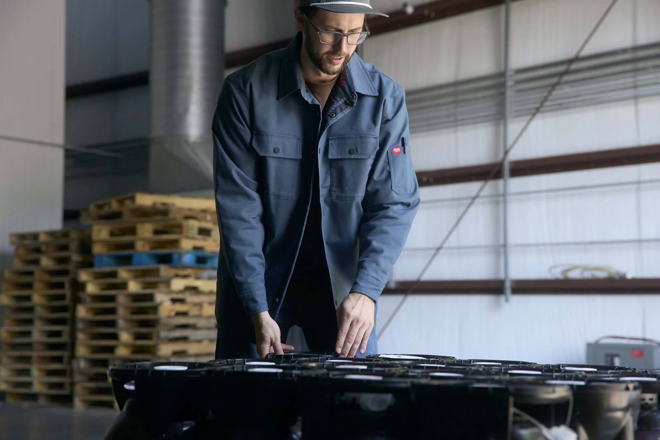 A man wearing glasses, a blue work jacket, and a cap is working with tools on a table inside a warehouse or industrial space, with wooden pallets stacked in the background.