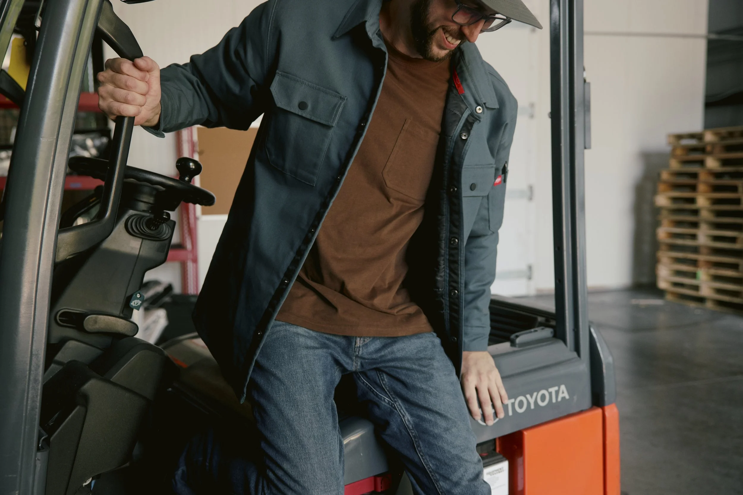 A man smiling and sitting inside a forklift, holding onto a metal bar. He is wearing a gray hat, a dark blue jacket, a brown shirt, and jeans. The forklift has a visible steering wheel and controls, and the background includes wooden pallets and a ga