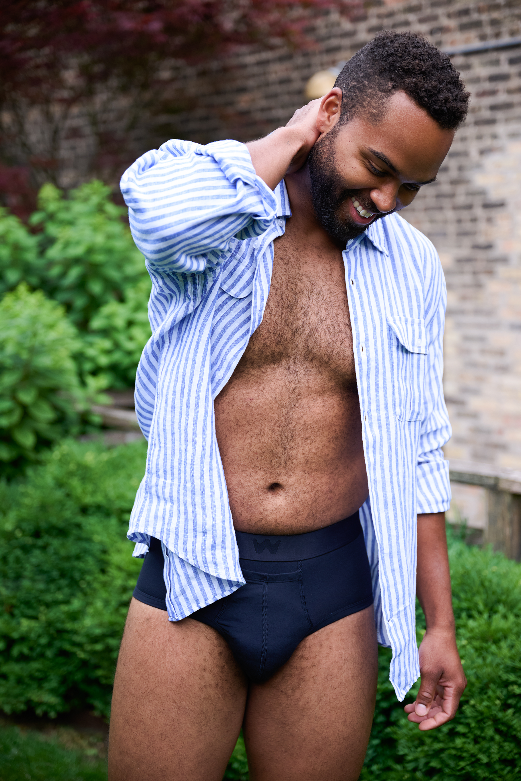 A man with dark curly hair and a beard is smiling outdoors in front of a brick wall and greenery. He wears an unbuttoned blue-and-white striped shirt, revealing his chest and abdomen, and black underwear. His right hand is touching his neck.