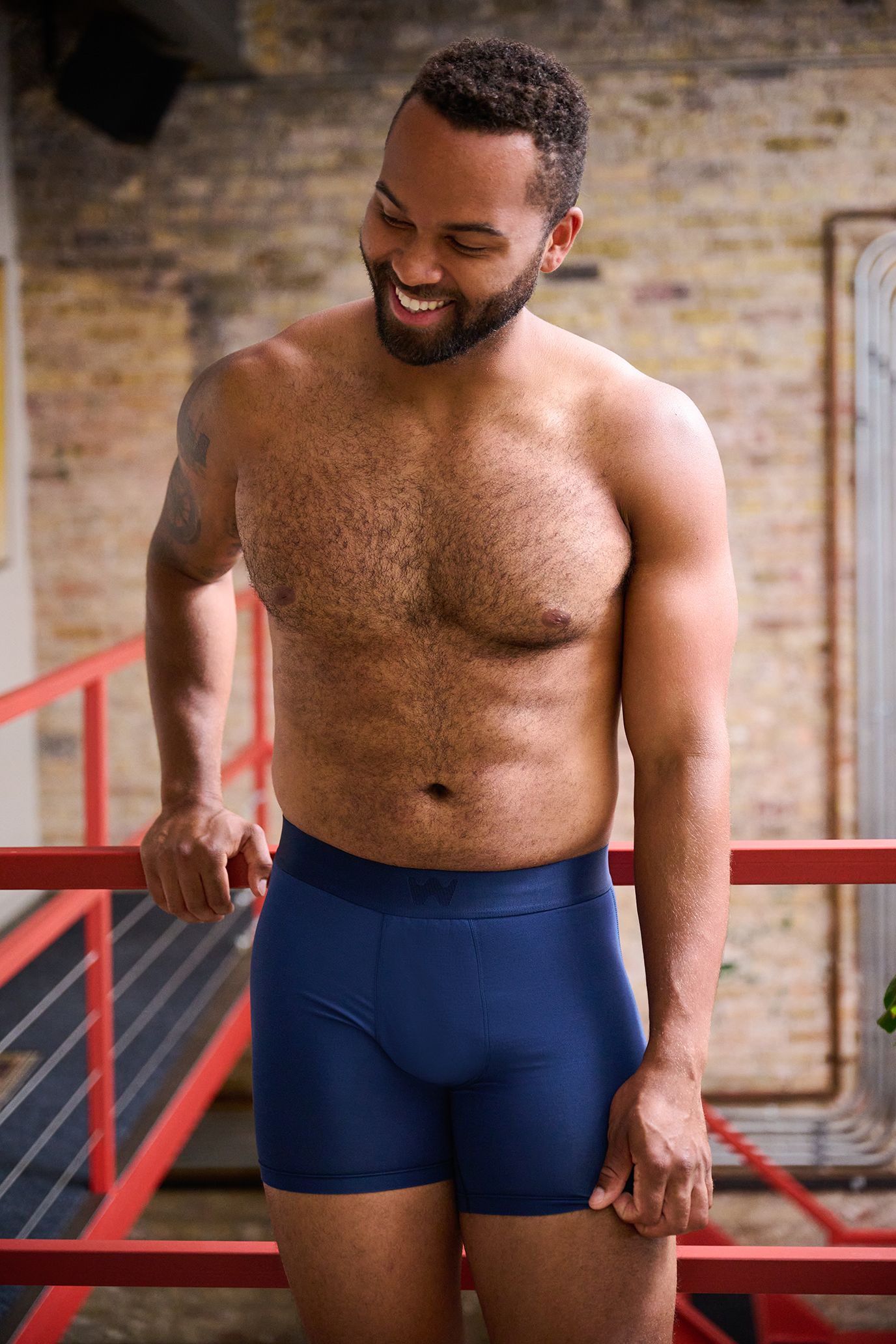 A shirtless man with a beard is smiling while standing on a gym staircase. He is wearing blue shorts and has tattoos on his left arm. The background features a exposed brick wall and gym equipment.