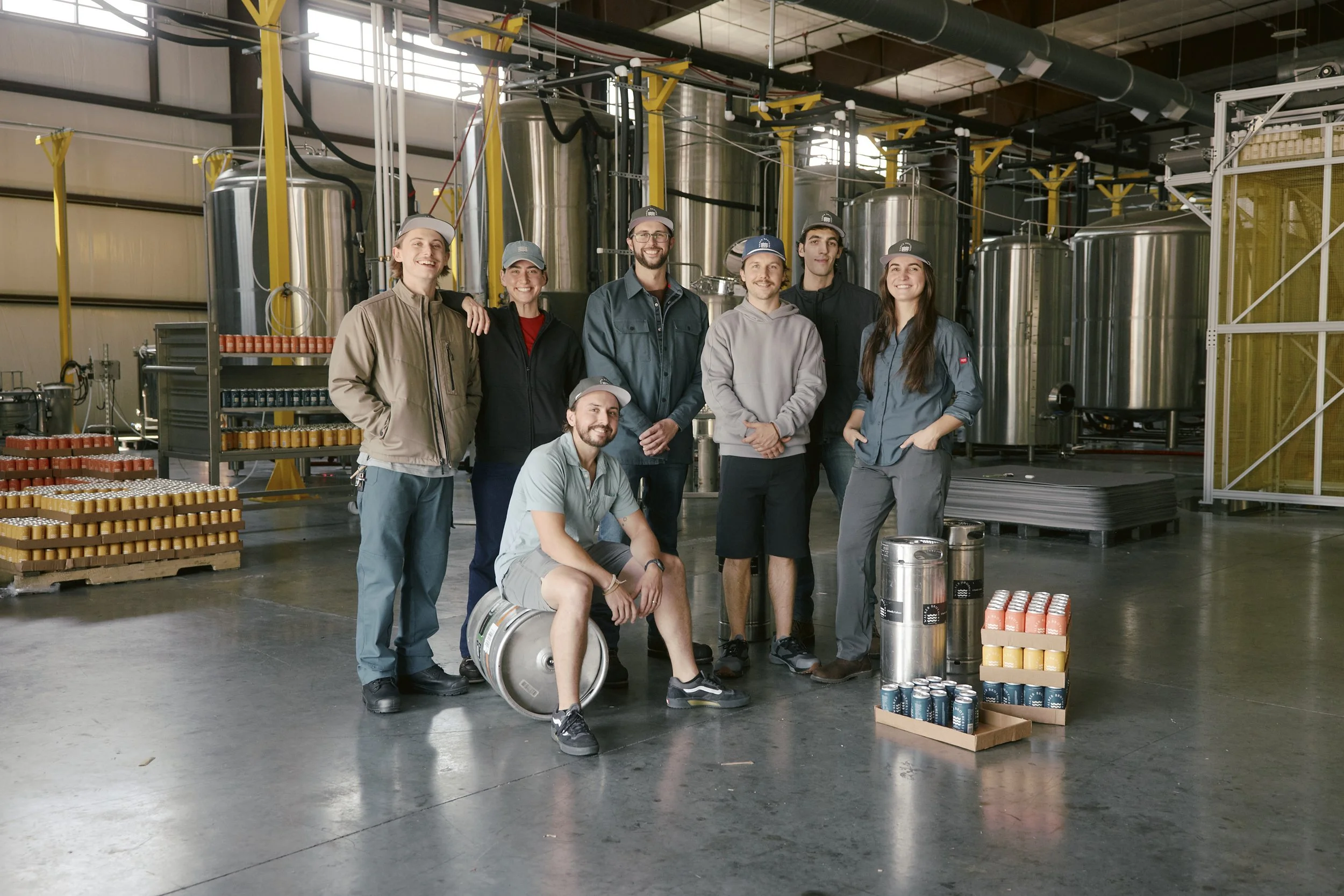 Group of eight smiling people in casual work clothes and caps in an industrial brewery setting, with large stainless steel tanks and equipment behind them, some standing and two sitting or kneeling with a metal barrel and cans of brewing ingredients 