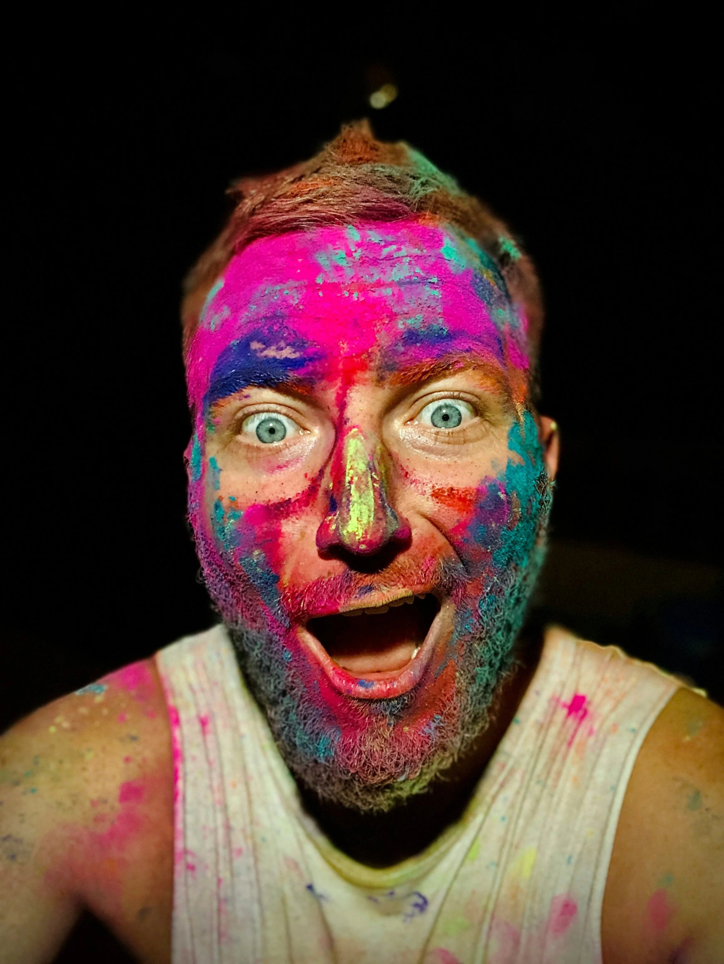 A person with blue eyes and facial hair, covered in bright colored powders including pink, blue, yellow, and green, celebrating during a festival with a joyful expression and mouth open.