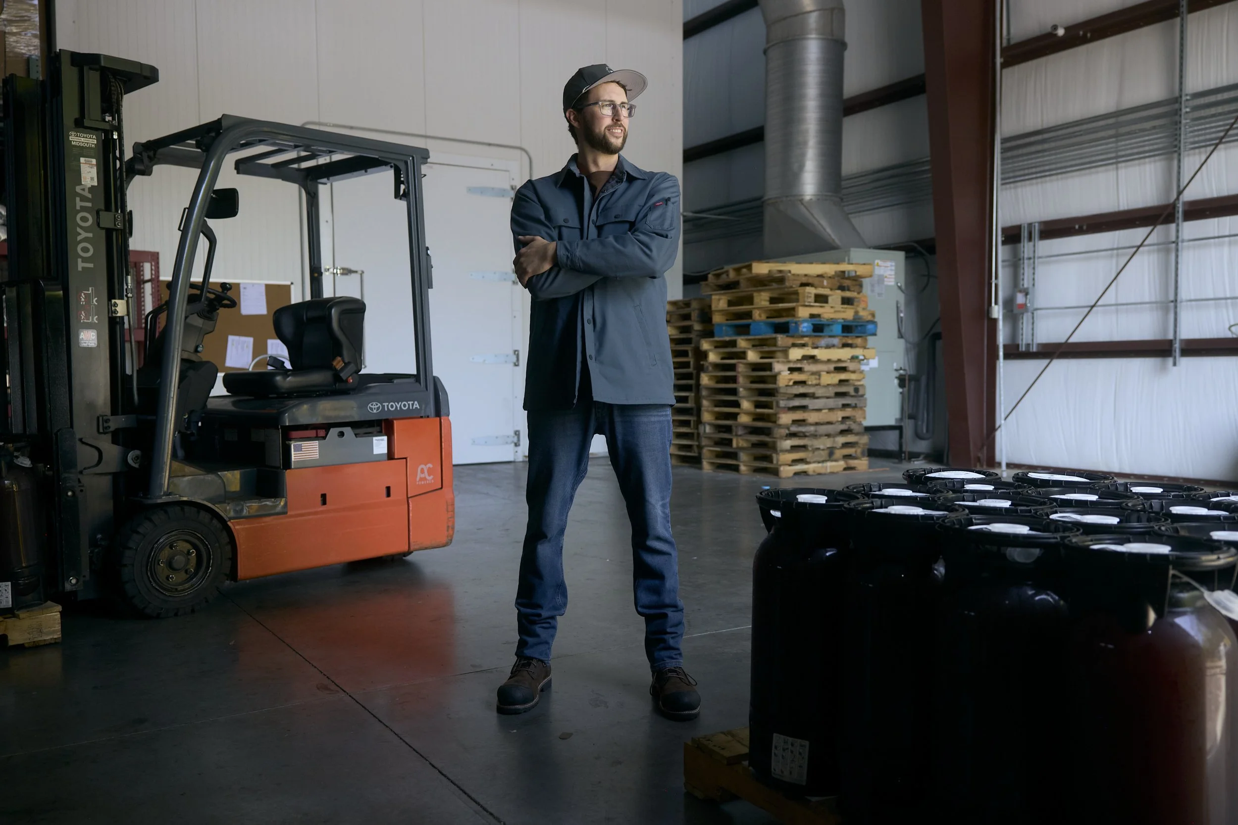 A man with glasses and a beard standing in an industrial warehouse. He is wearing a dark jacket, jeans, and a cap with arms crossed. To his left is an orange Toyota forklift, and behind him are stacked wooden pallets and containers of liquids on the 