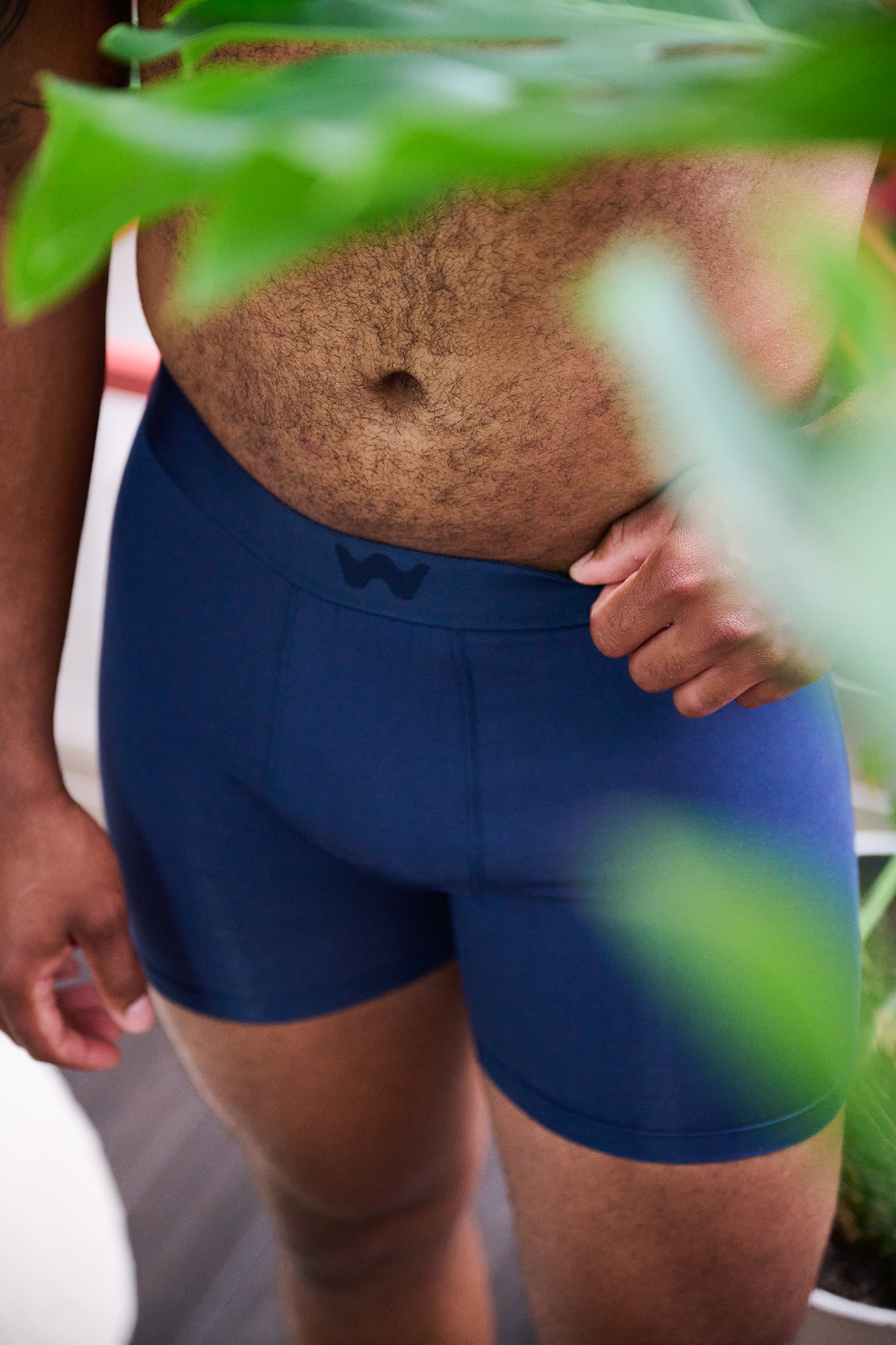 Close-up of a person's midsection in blue athletic shorts, with one hand gripping the waistband near the hip, seen through green leaves in the foreground.