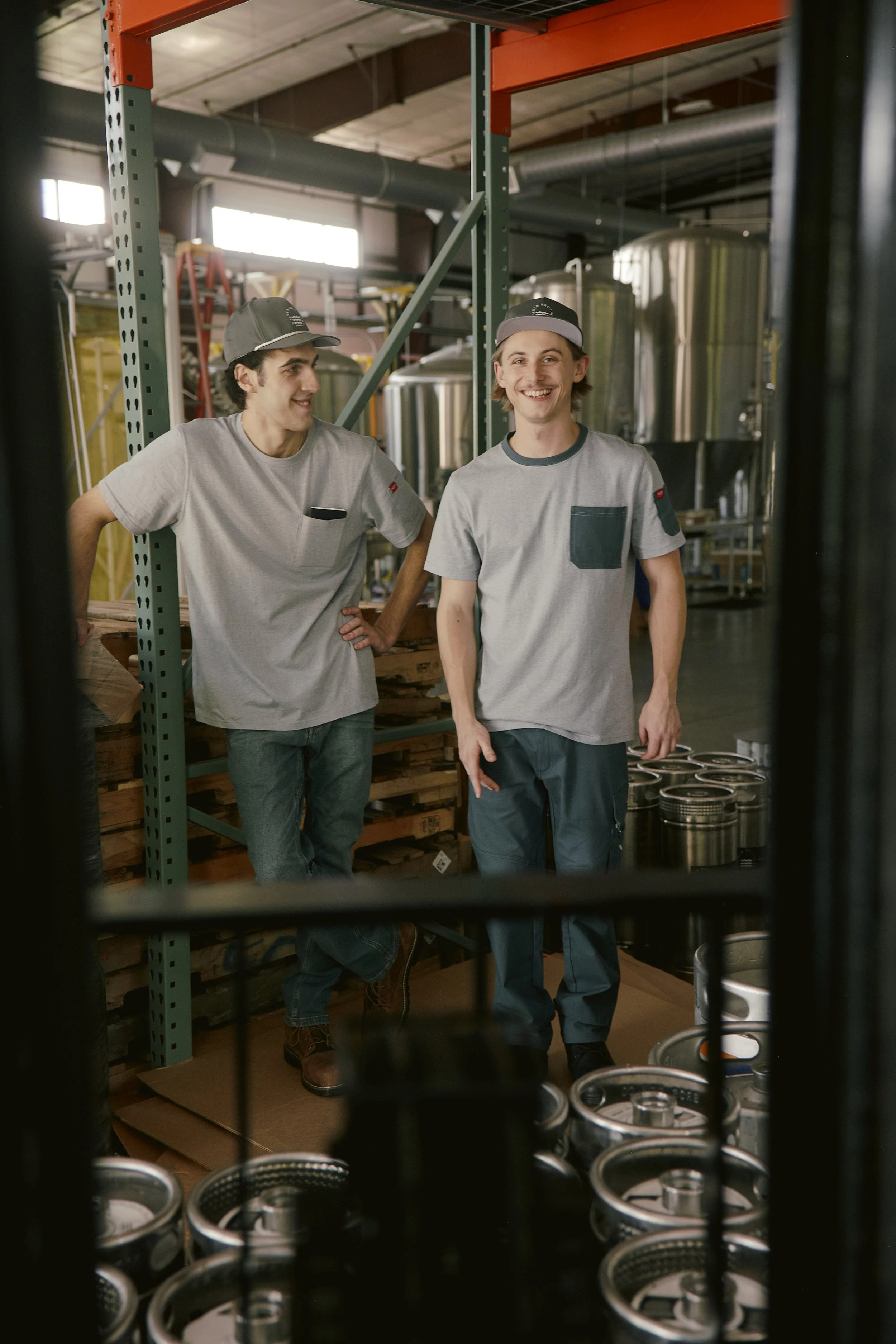 Two young men in casual work attire, wearing hats, standing inside a brewery or cidery with stainless steel fermentation tanks and shelves with supplies.